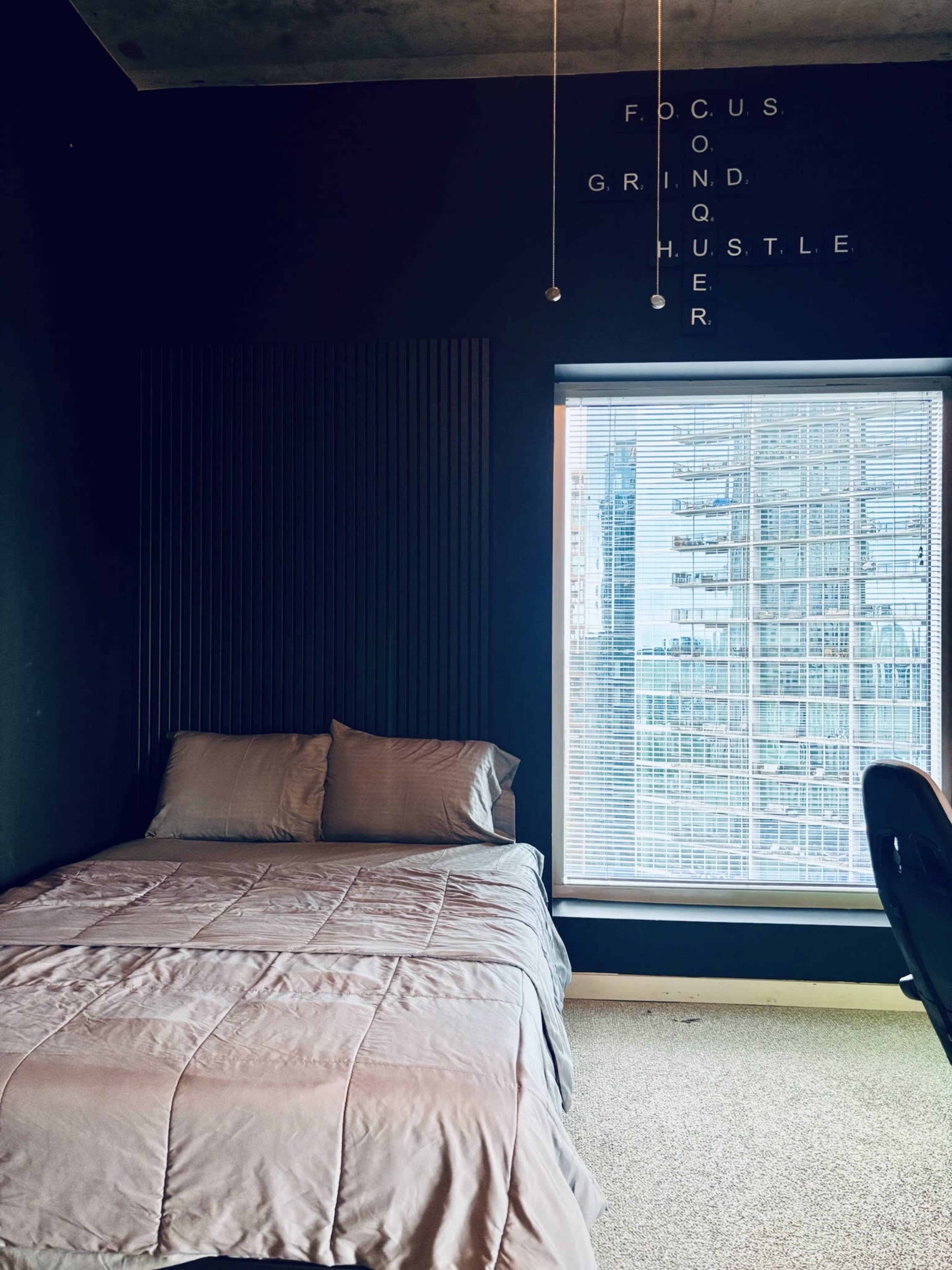 A neatly made bed with gray bedding is positioned next to a large window that reveals a view of tall buildings.
