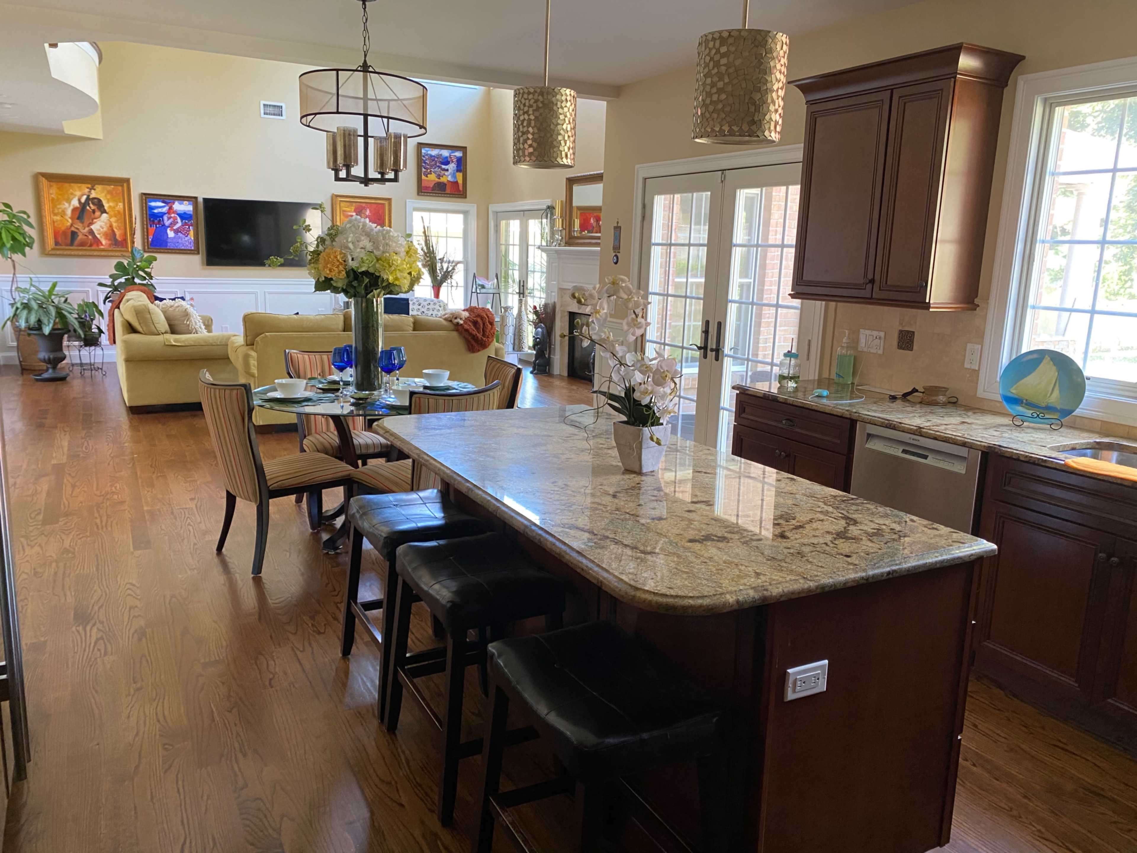 A modern kitchen with a granite island, bar stools, and an open layout leading into a living area with large windows and colorful artwork.