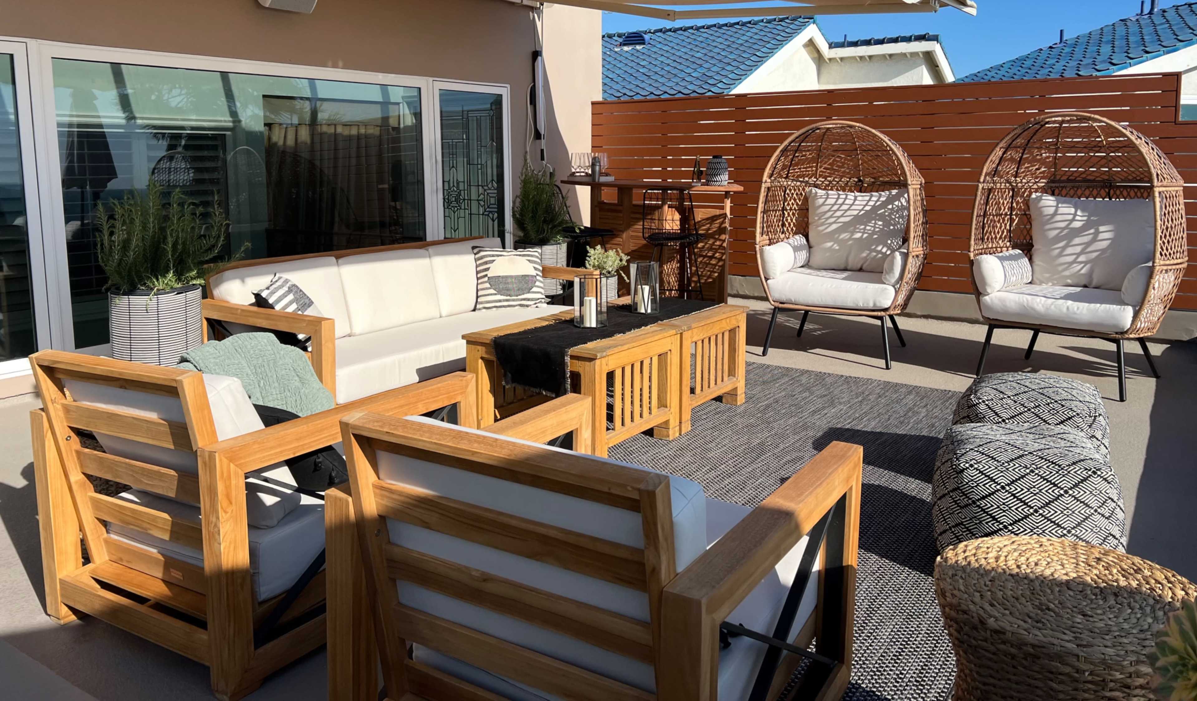 A modern outdoor patio setup featuring wooden furniture, including a couch and two hanging chairs, arranged around a central coffee table on a carpet.