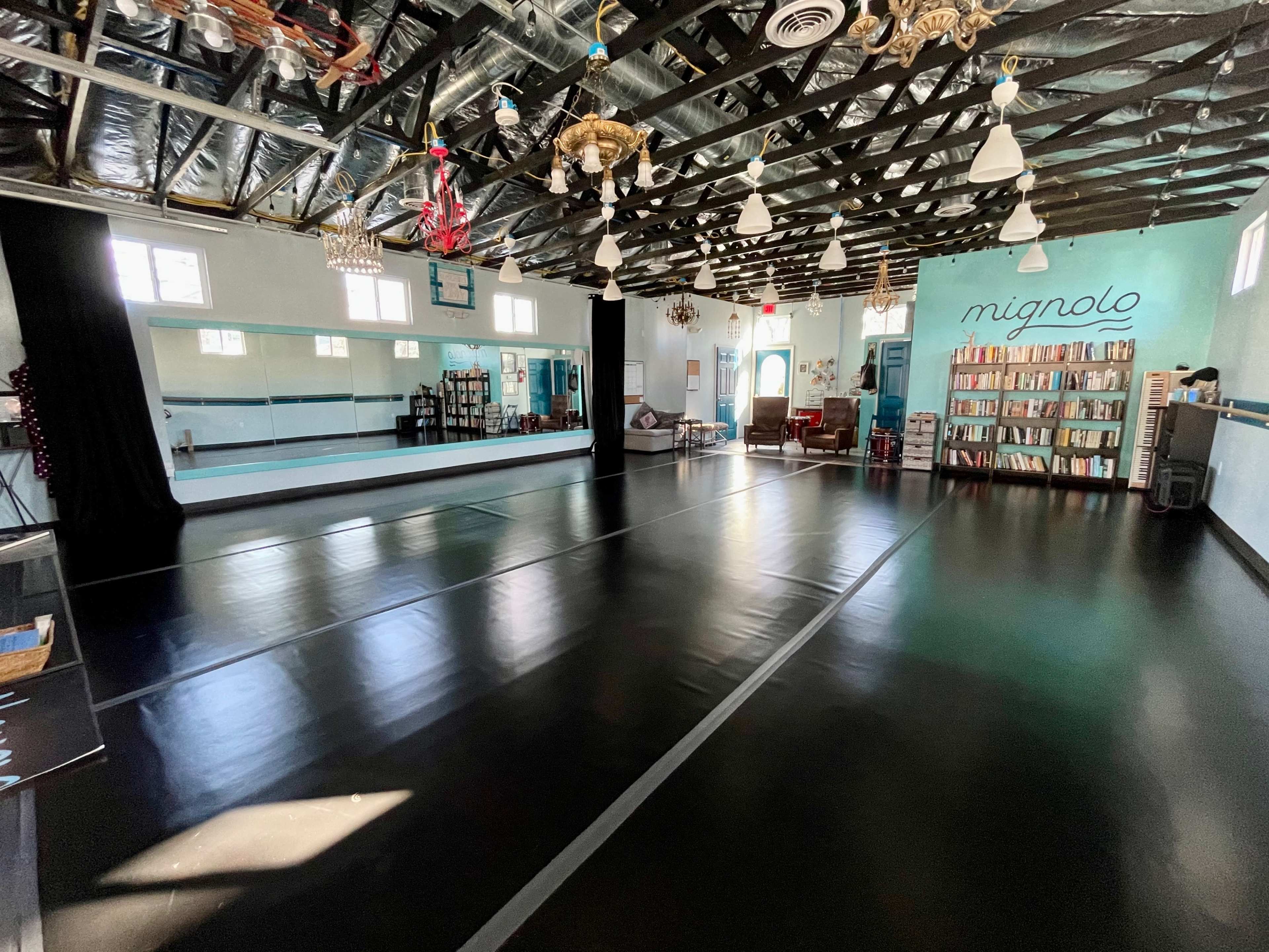 A spacious dance studio with black flooring, large mirrors, and bookshelves along one wall.