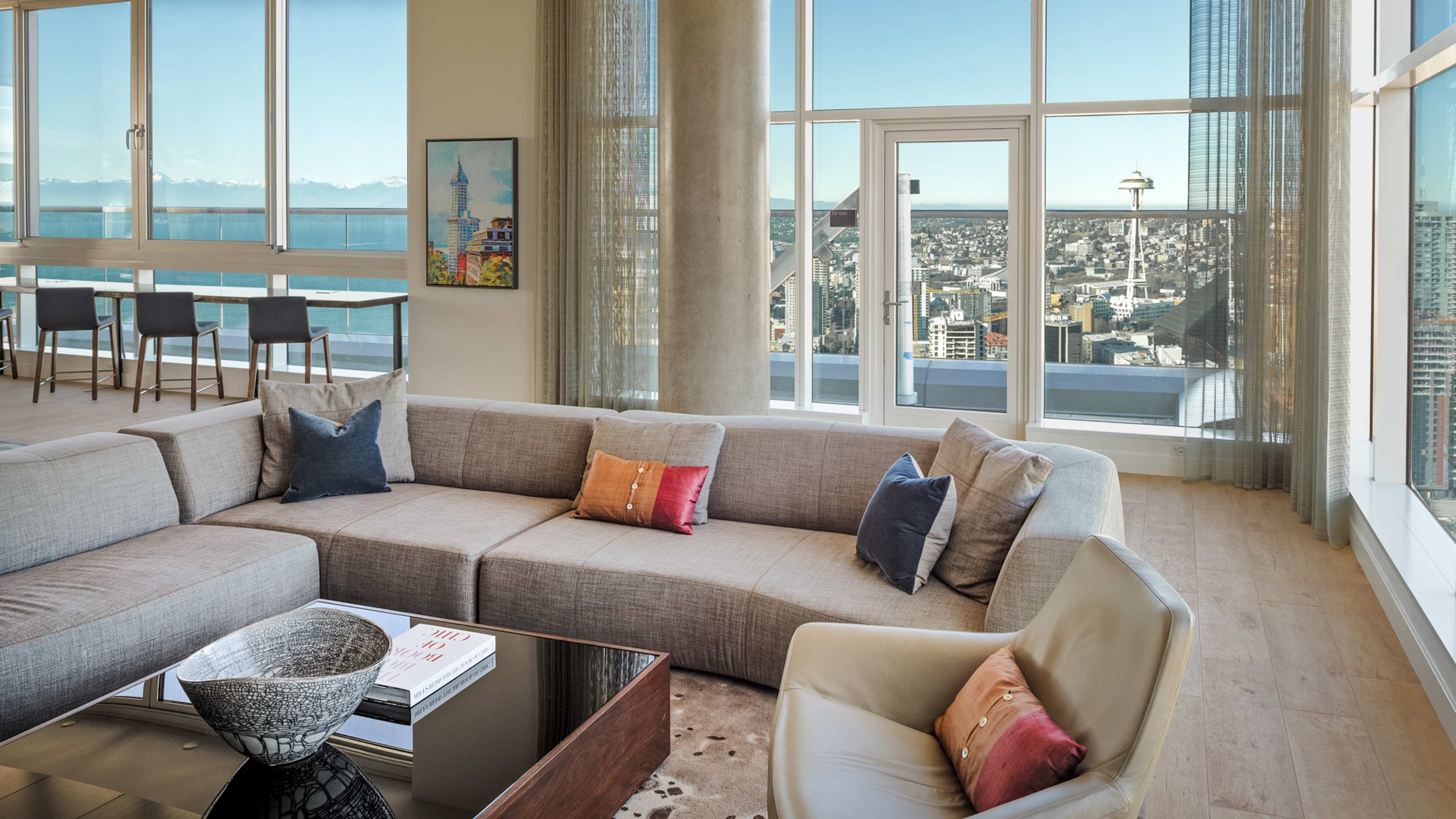 A modern living room with a large sectional sofa, glass table, and expansive windows offering a panoramic view of a city skyline and mountains in the distance.