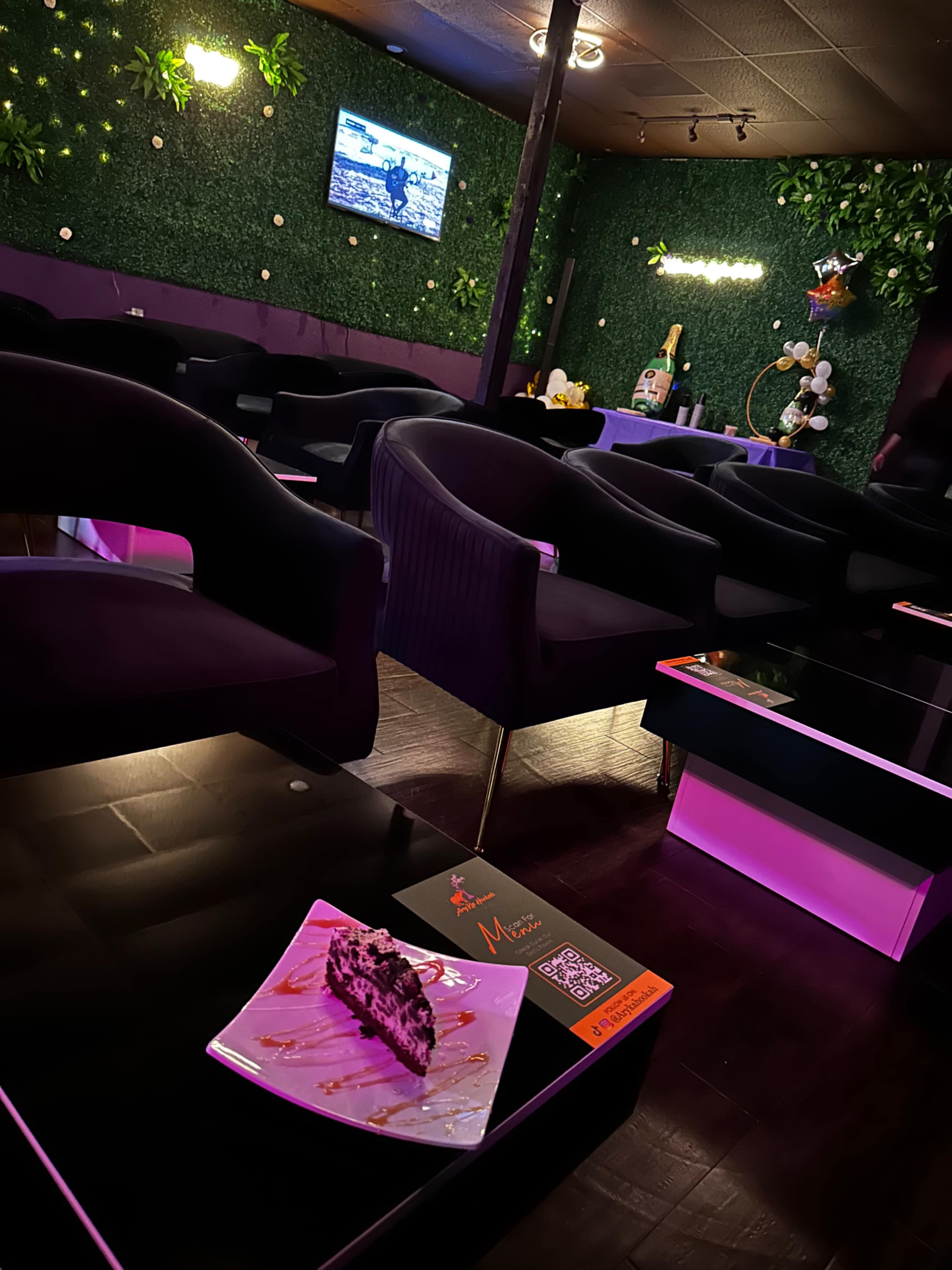 A cozy lounge area features dark upholstered chairs, a dessert on a plate, and a television mounted on a wall adorned with greenery.