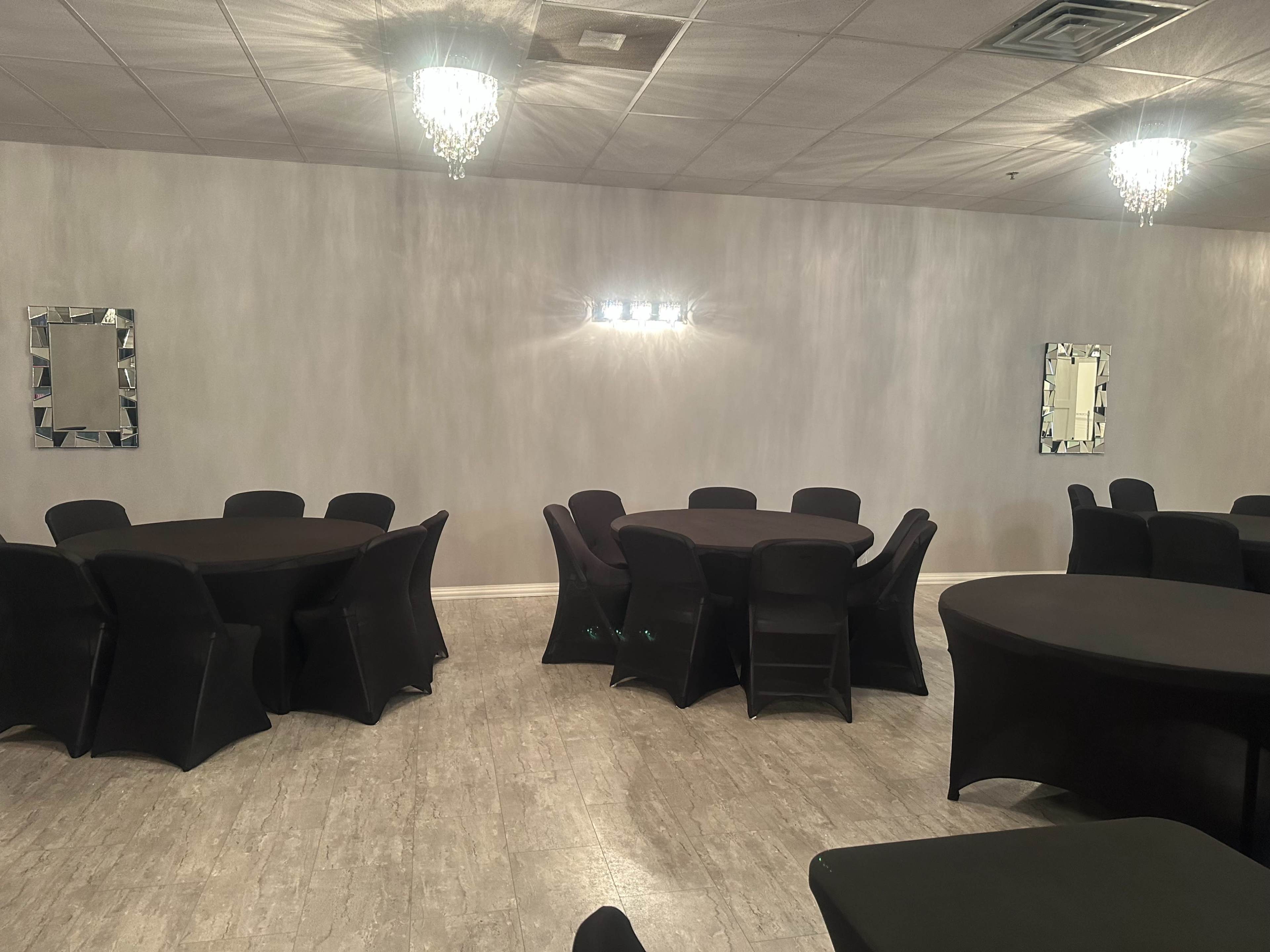 A spacious event room with several round black tables and chairs, illuminated by wall-mounted lights and crystal chandeliers.