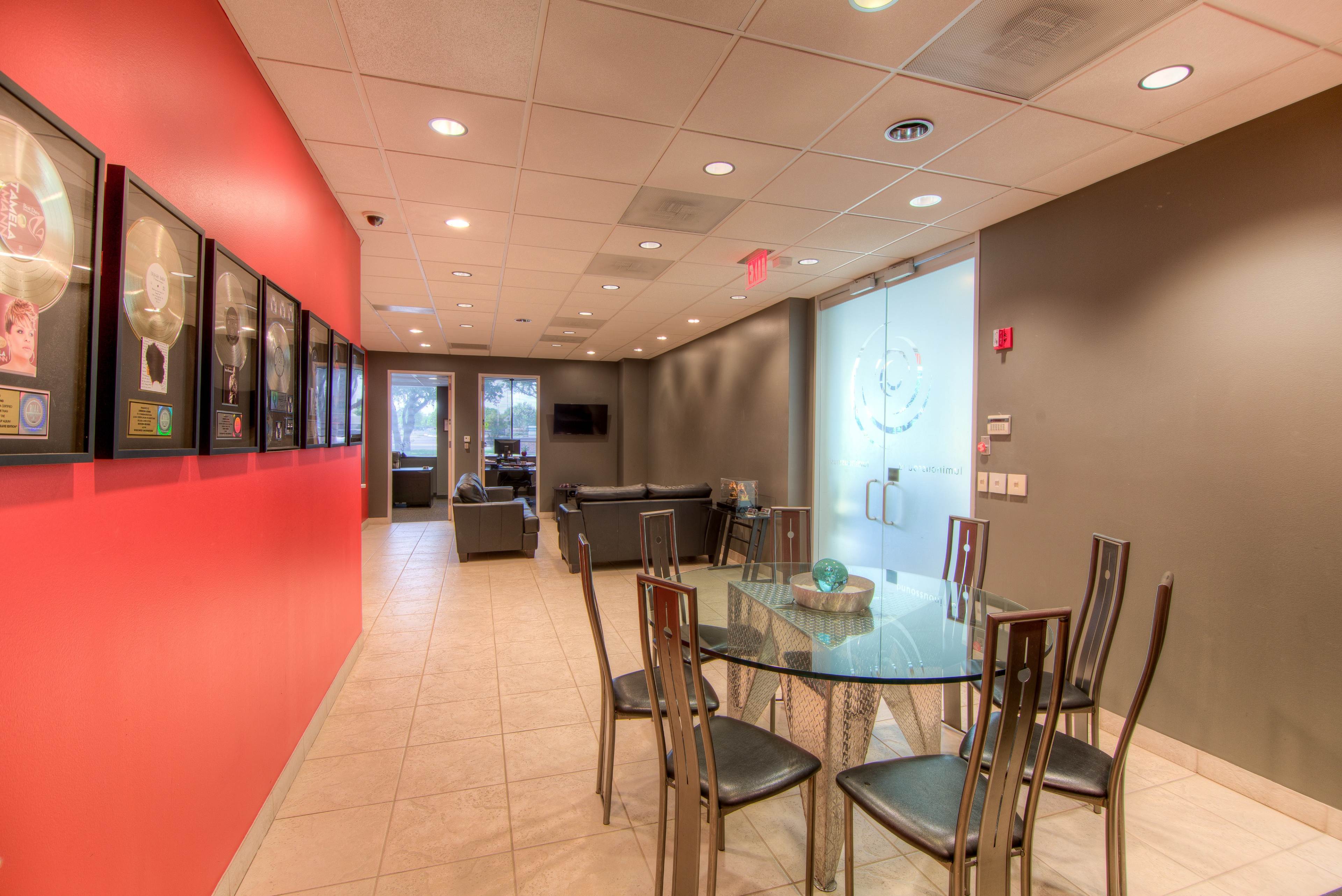 The image shows a hallway that leads to a lounge area with a glass-topped dining table and black chairs, framed awards on the wall, and a couch in the background.