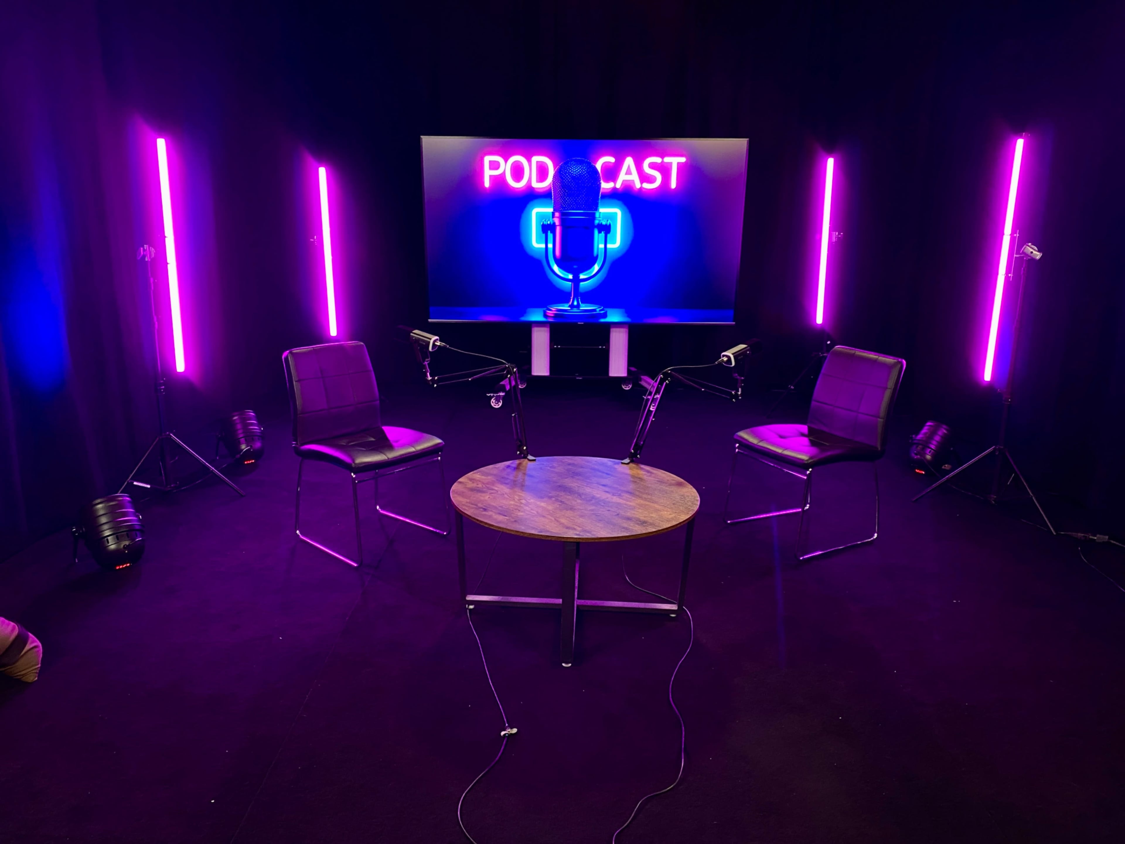 The image shows a podcast studio setup with two chairs, a round table, and a background display featuring a microphone graphic illuminated by purple lights.