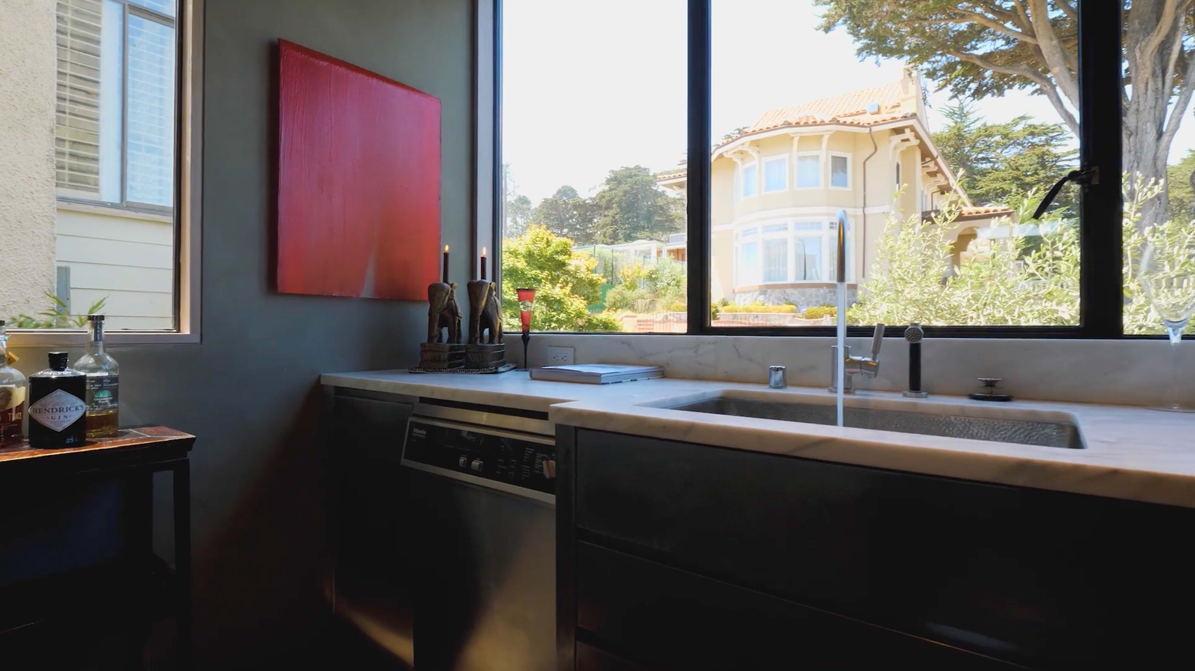A kitchen with a sink and countertop overlooks a garden and a large house through a large window.