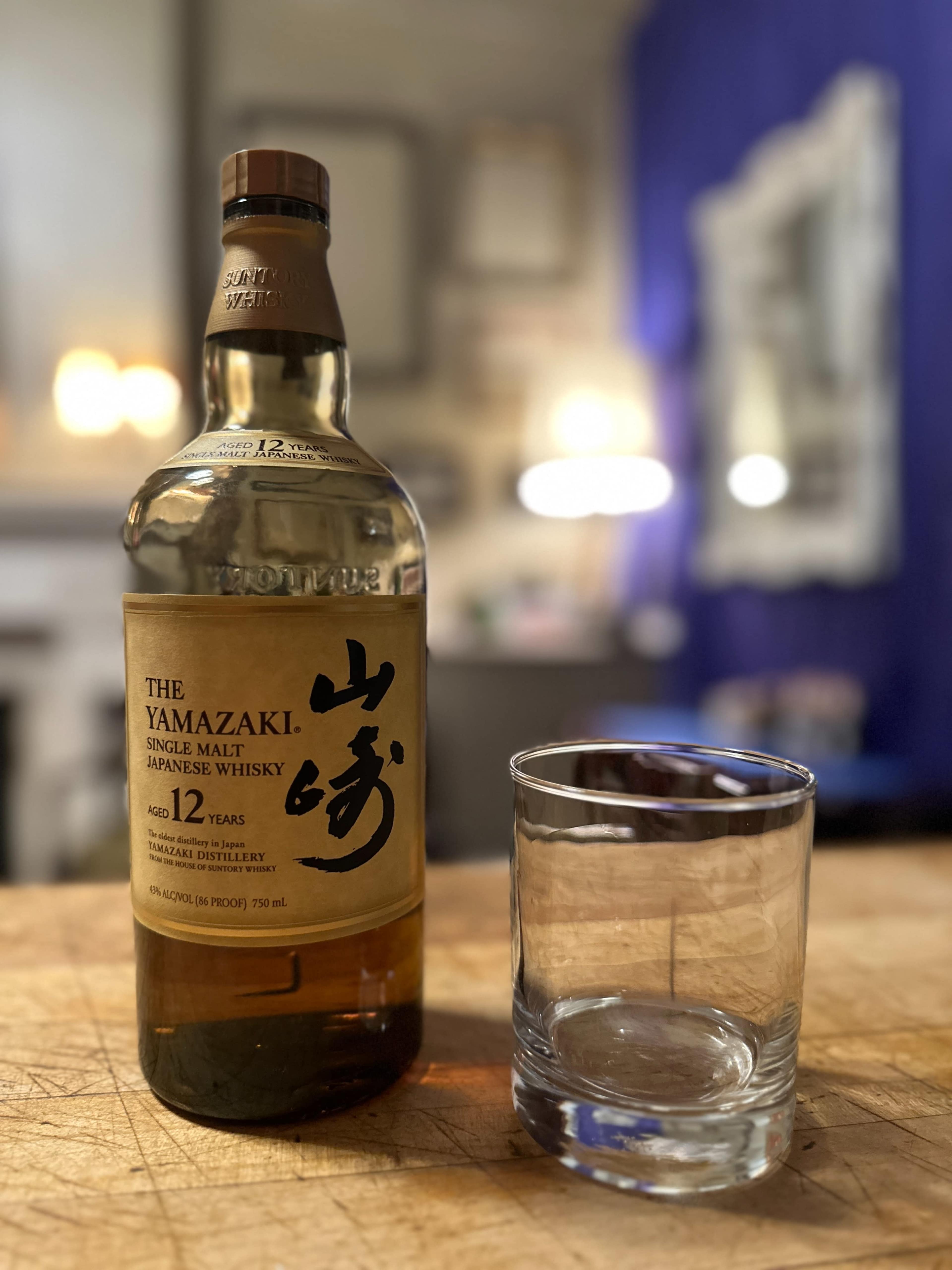 A bottle of Yamazaki 12-Year-Old Single Malt Japanese Whisky sits on a wooden surface next to a short, clear glass.