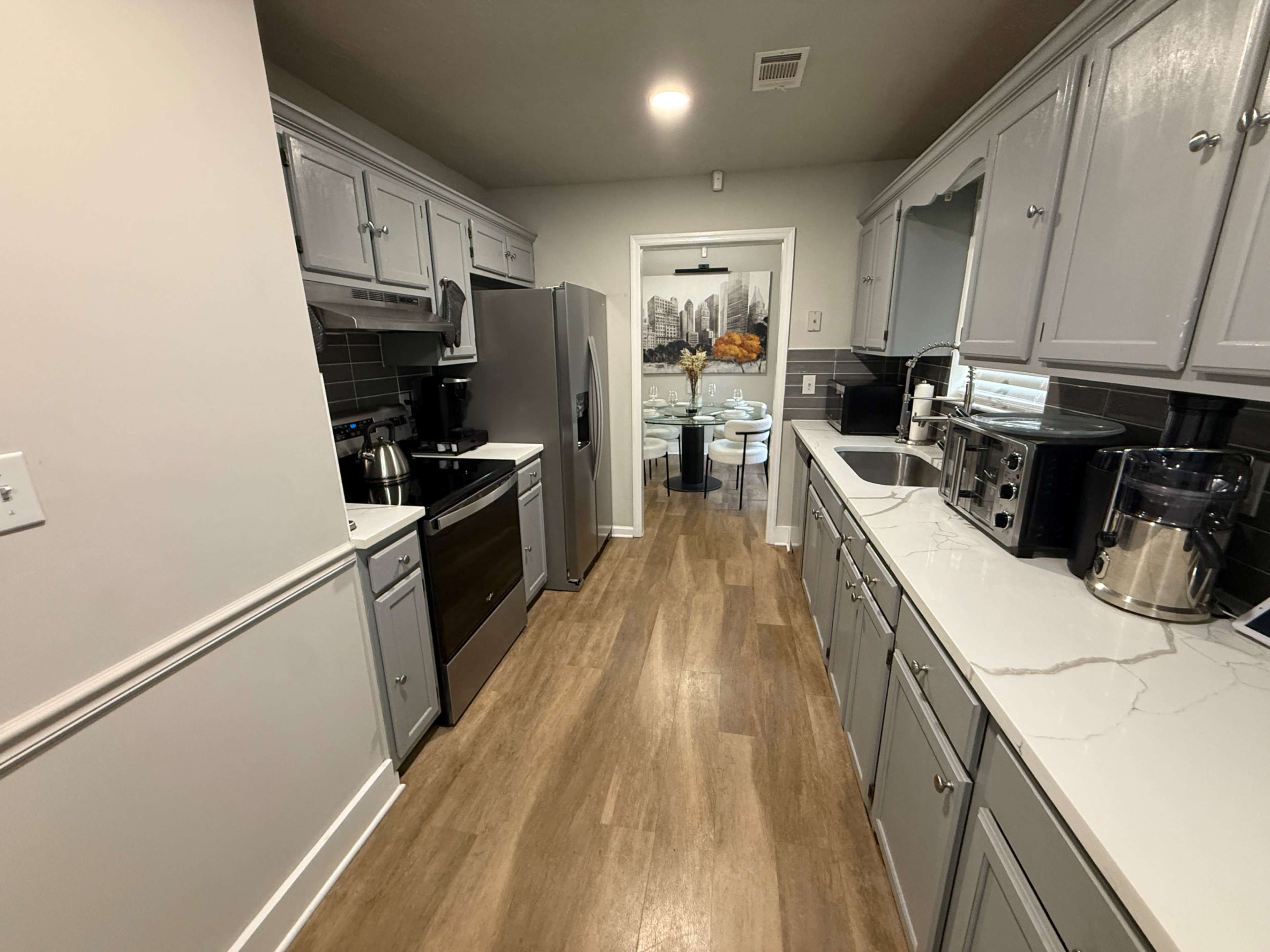 The image shows a modern kitchen with gray cabinetry, stainless steel appliances, and a light-colored countertop, leading to a dining area visible in the background.