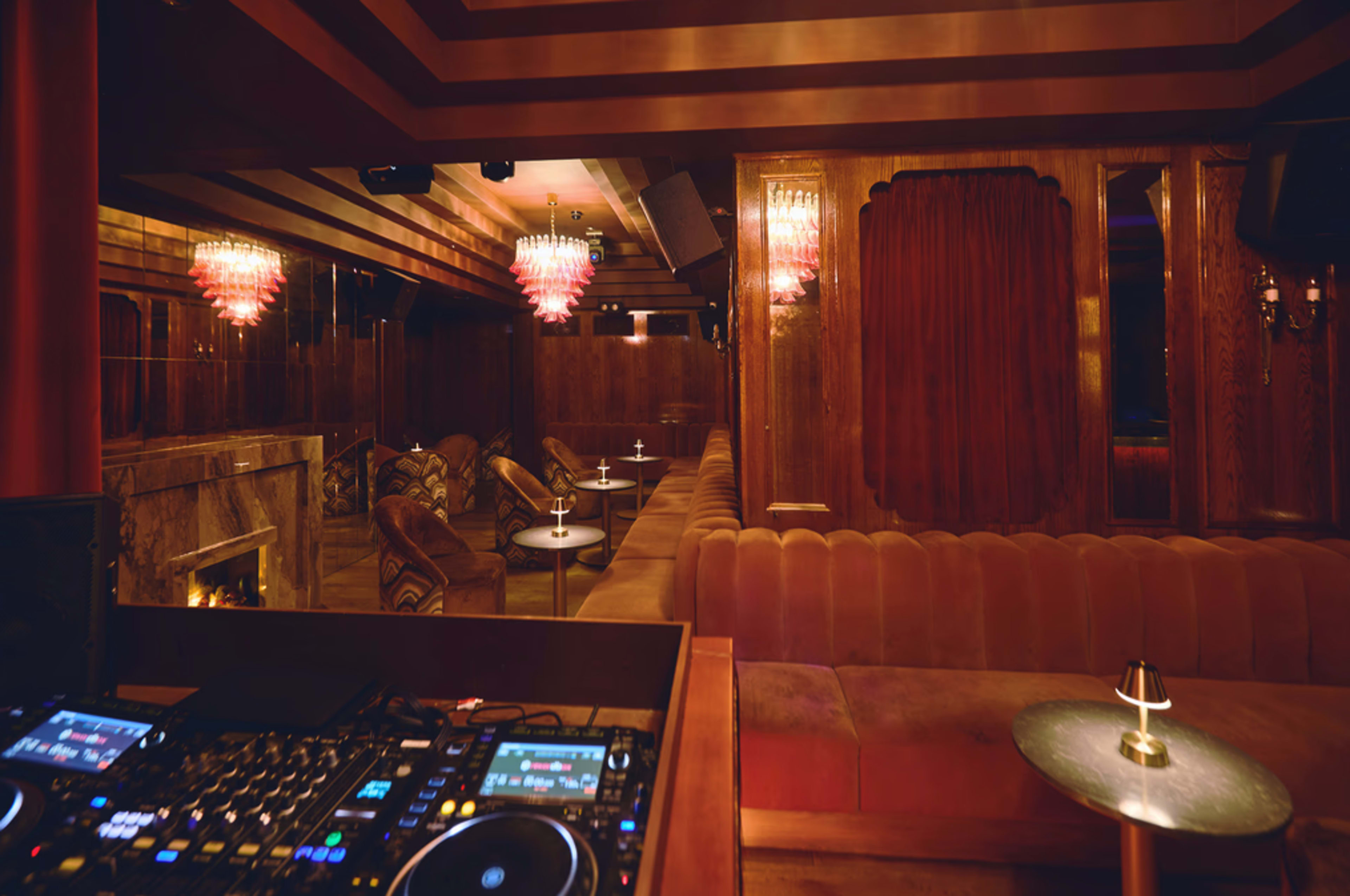 The image shows a dimly lit lounge area featuring plush seating, small tables, and a DJ setup in the foreground.
