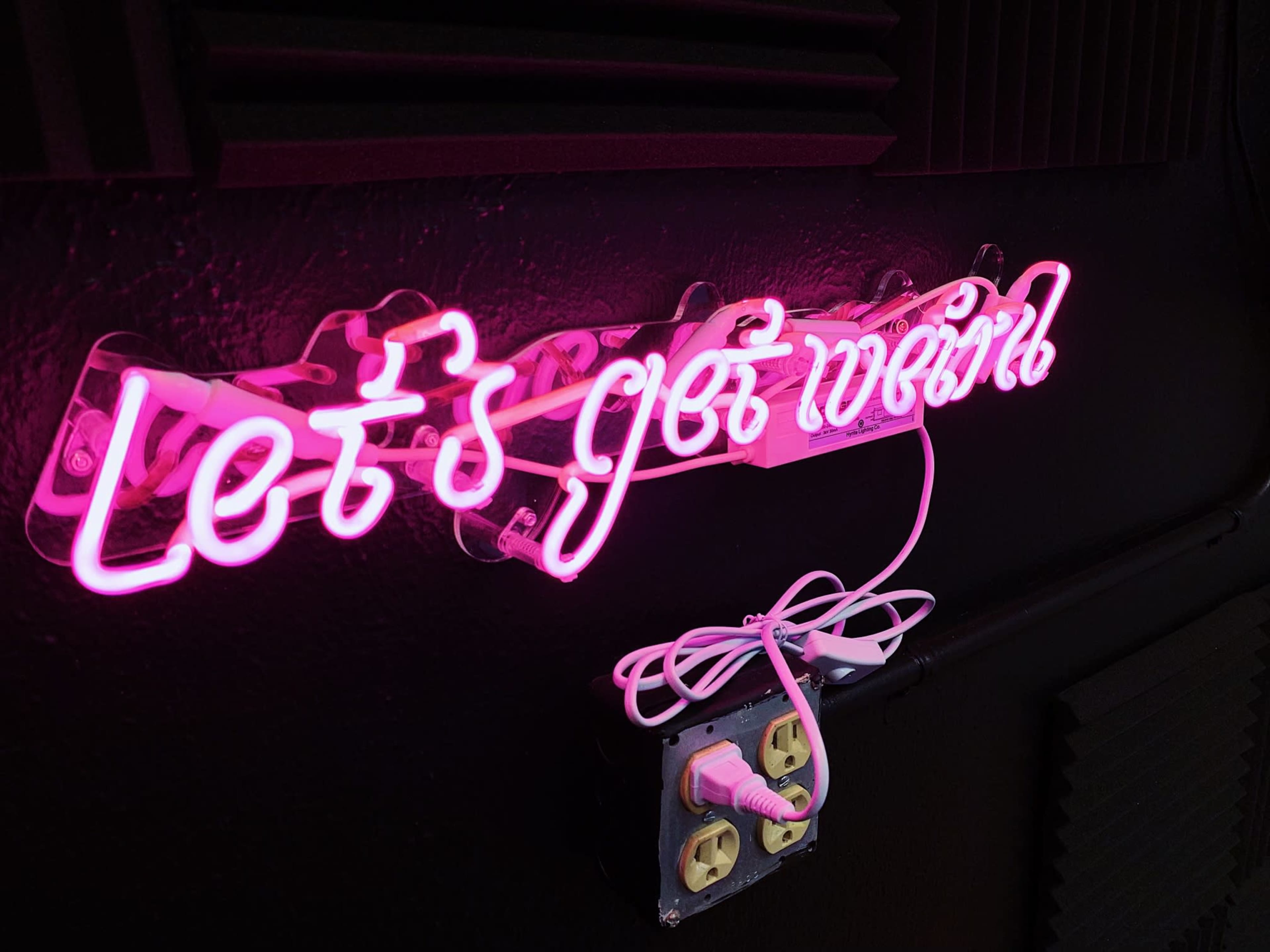 A neon sign reading "Let's get weird" is mounted on a dark wall with a visible power outlet nearby.
