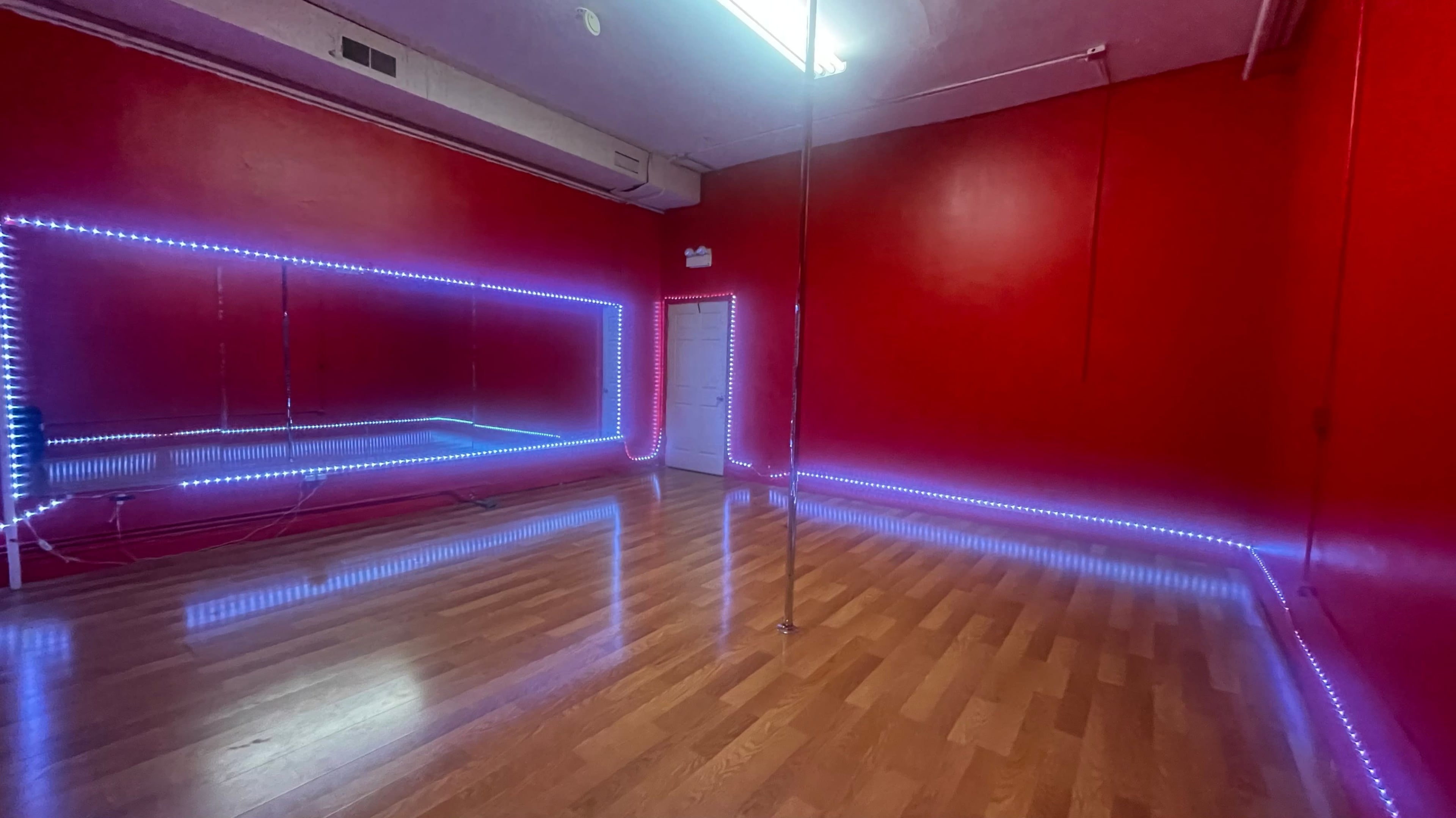 A dance studio with red walls, wooden flooring, and a dance pole illuminated by LED lights along the perimeter.
