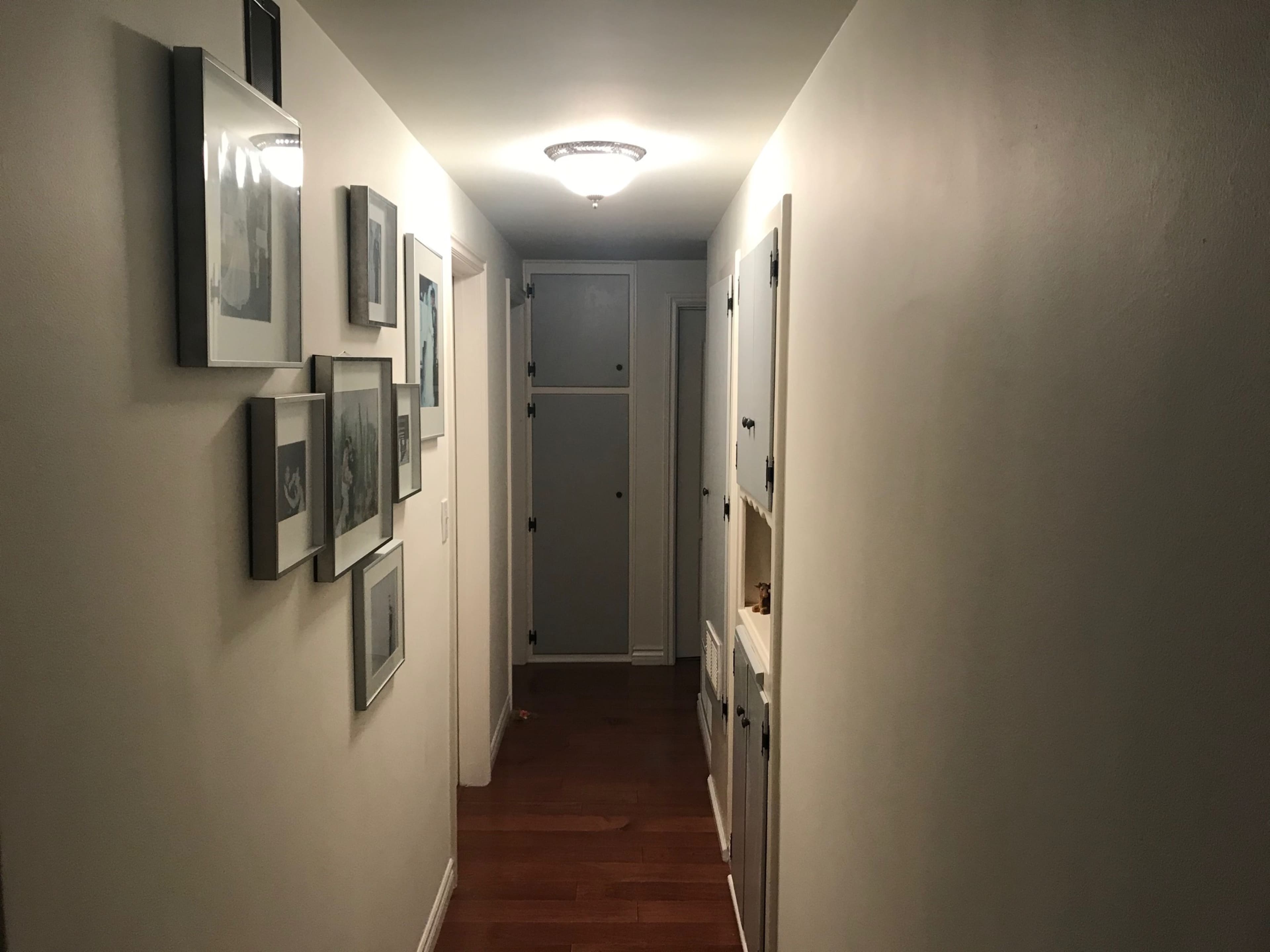 A narrow hallway with framed pictures on the left and closed doors on either side, leading to a bathroom at the end.