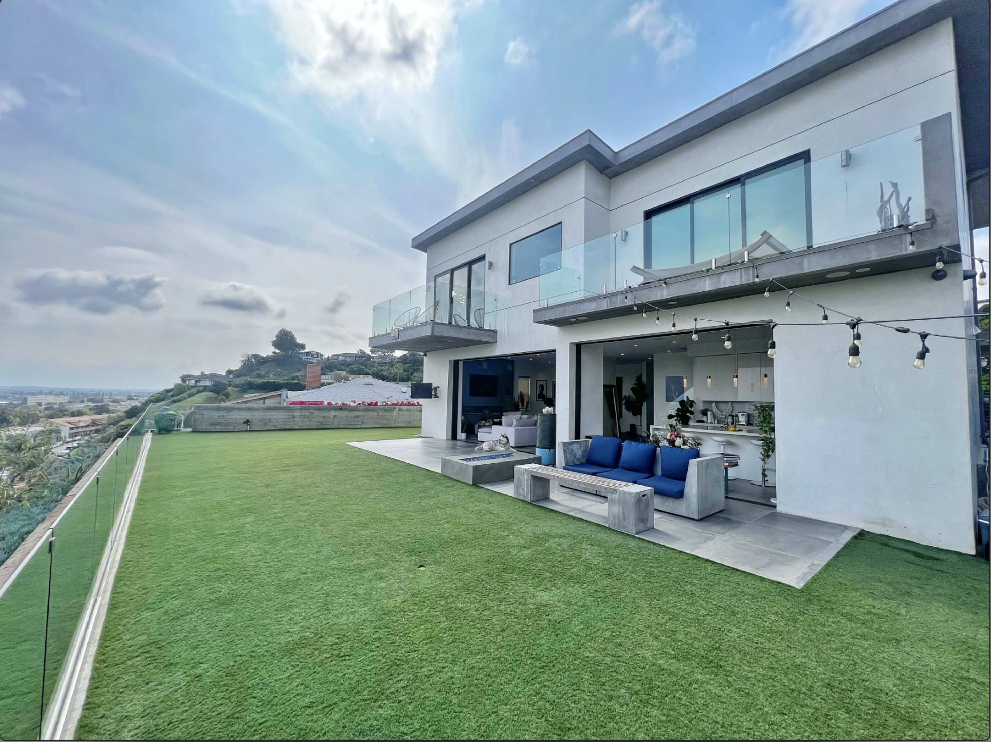 A modern two-story house features a terrace with glass railings, surrounded by a spacious green lawn and a cloudy sky.