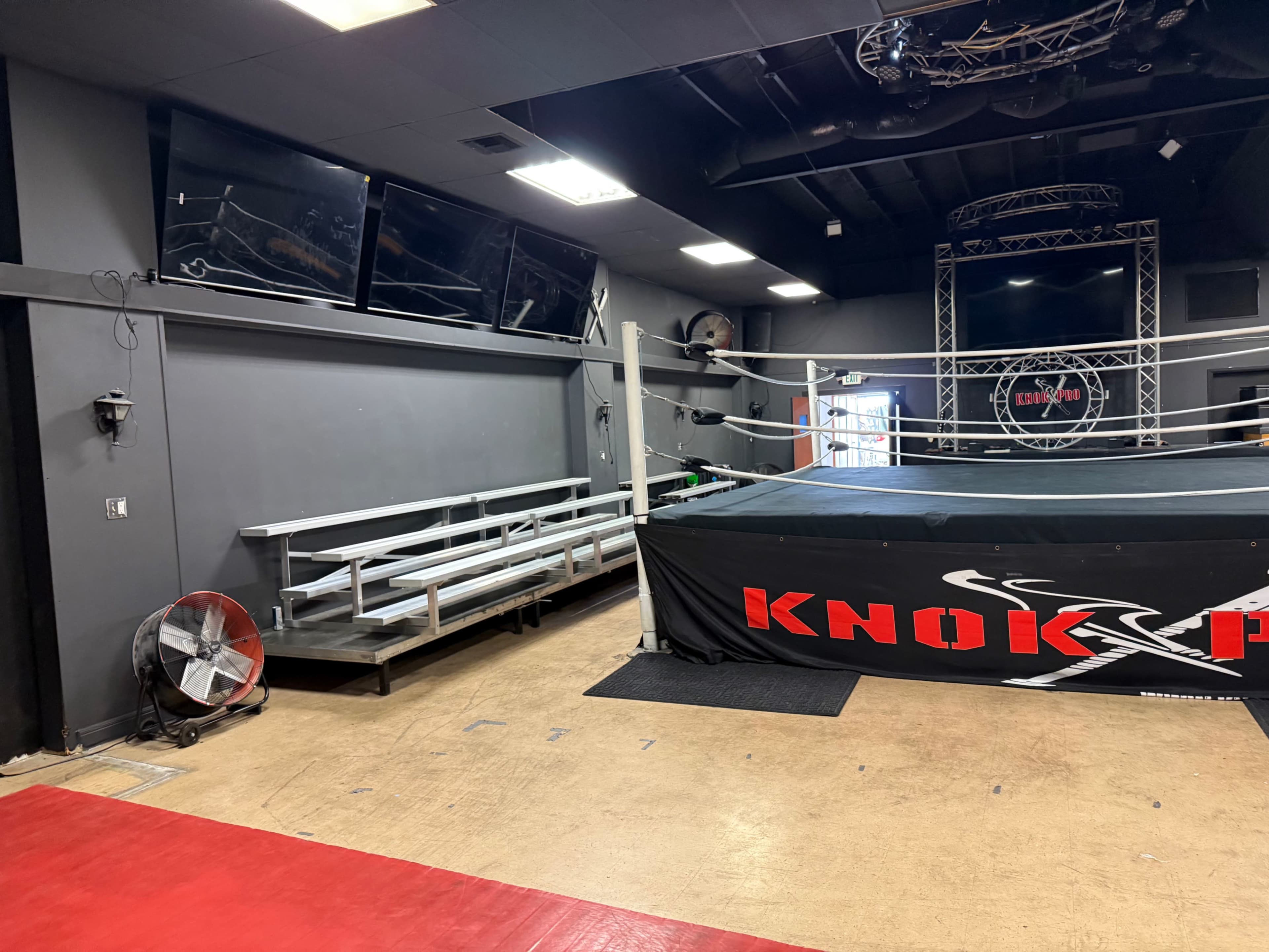 The image shows an indoor wrestling or boxing arena with a ring at the center, bleachers on one side, and a large fan nearby, all under industrial lighting.