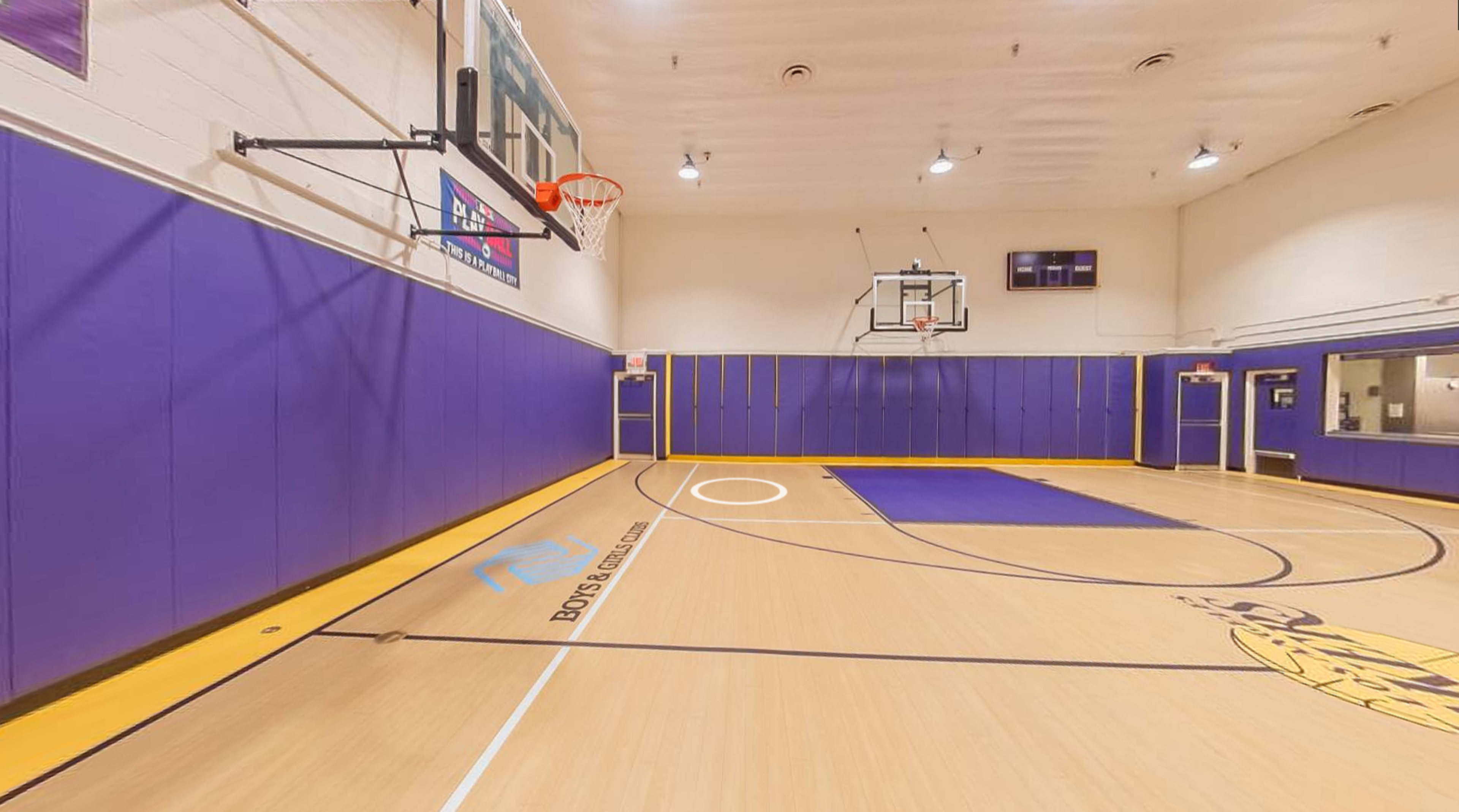A basketball gym with purple walls, a wooden floor featuring court markings, and two basketball hoops at either end.