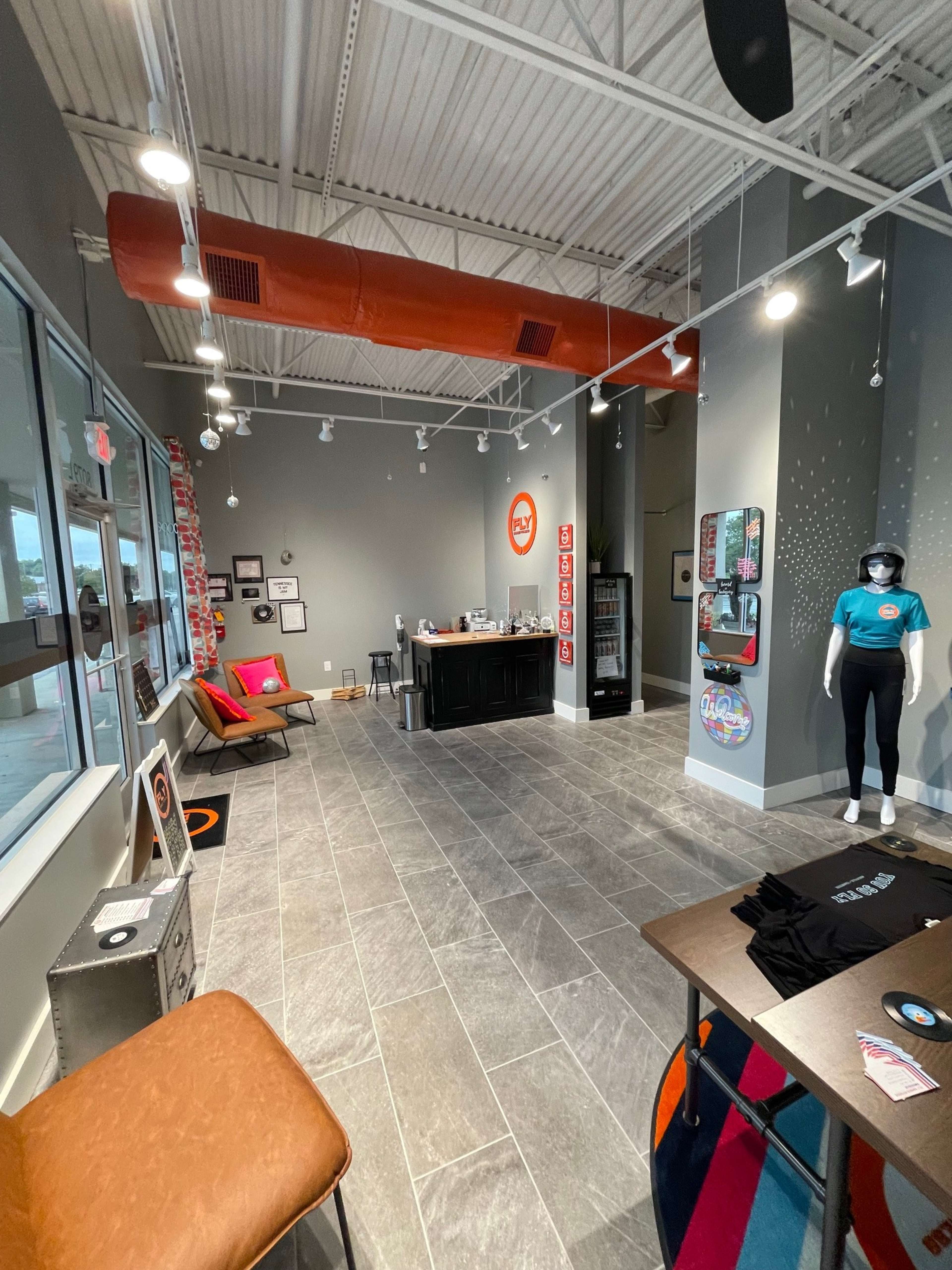 The image shows a modern retail space with gray walls, a reception counter, merchandise displays, seating areas, and large windows letting in natural light.