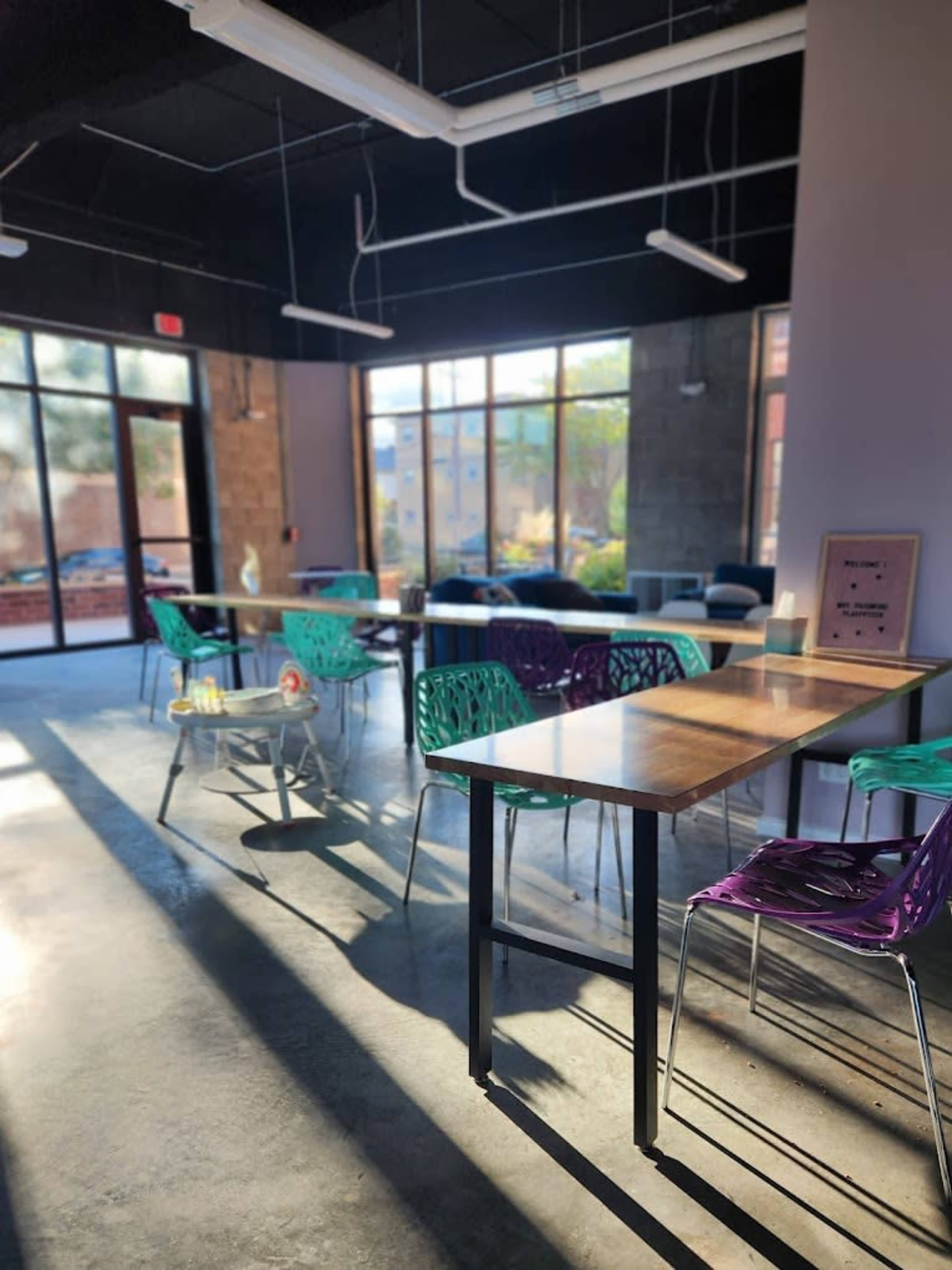 A well-lit, modern space features colorful chairs surrounding wooden tables, with large windows allowing natural light to fill the area.