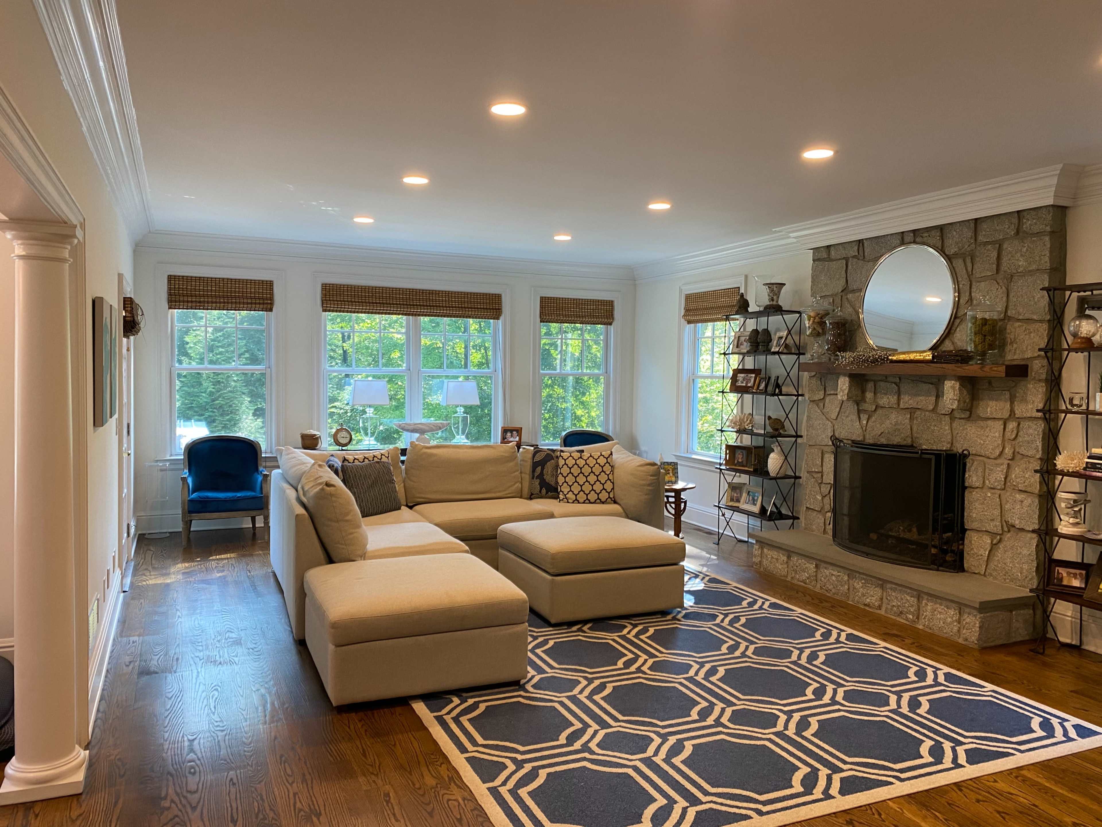 The living room features a large sectional sofa, a stone fireplace with a round mirror above it, and large windows allowing natural light to enter.