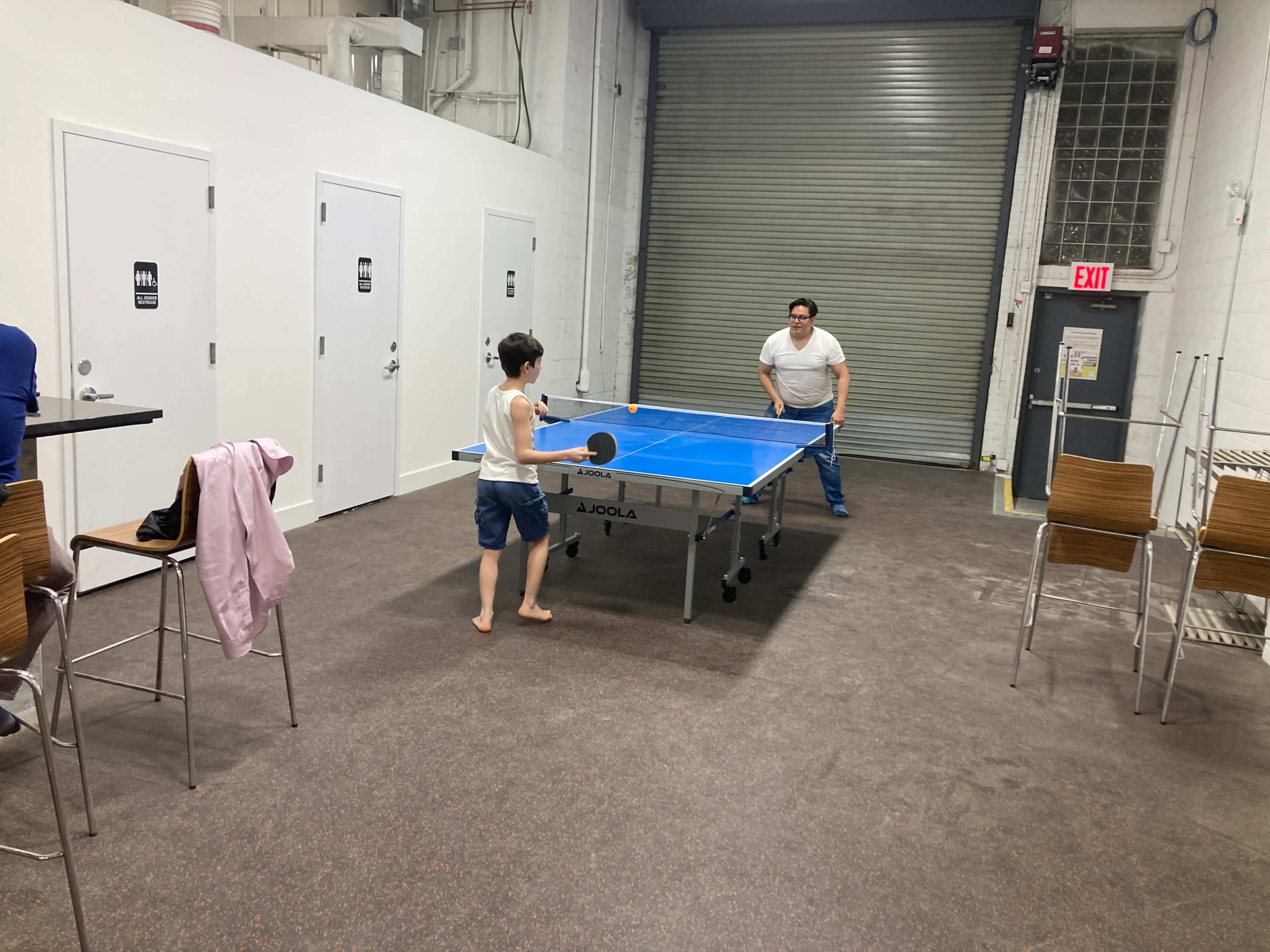 Two children are playing table tennis in a recreational room with a closed metal garage door in the background.