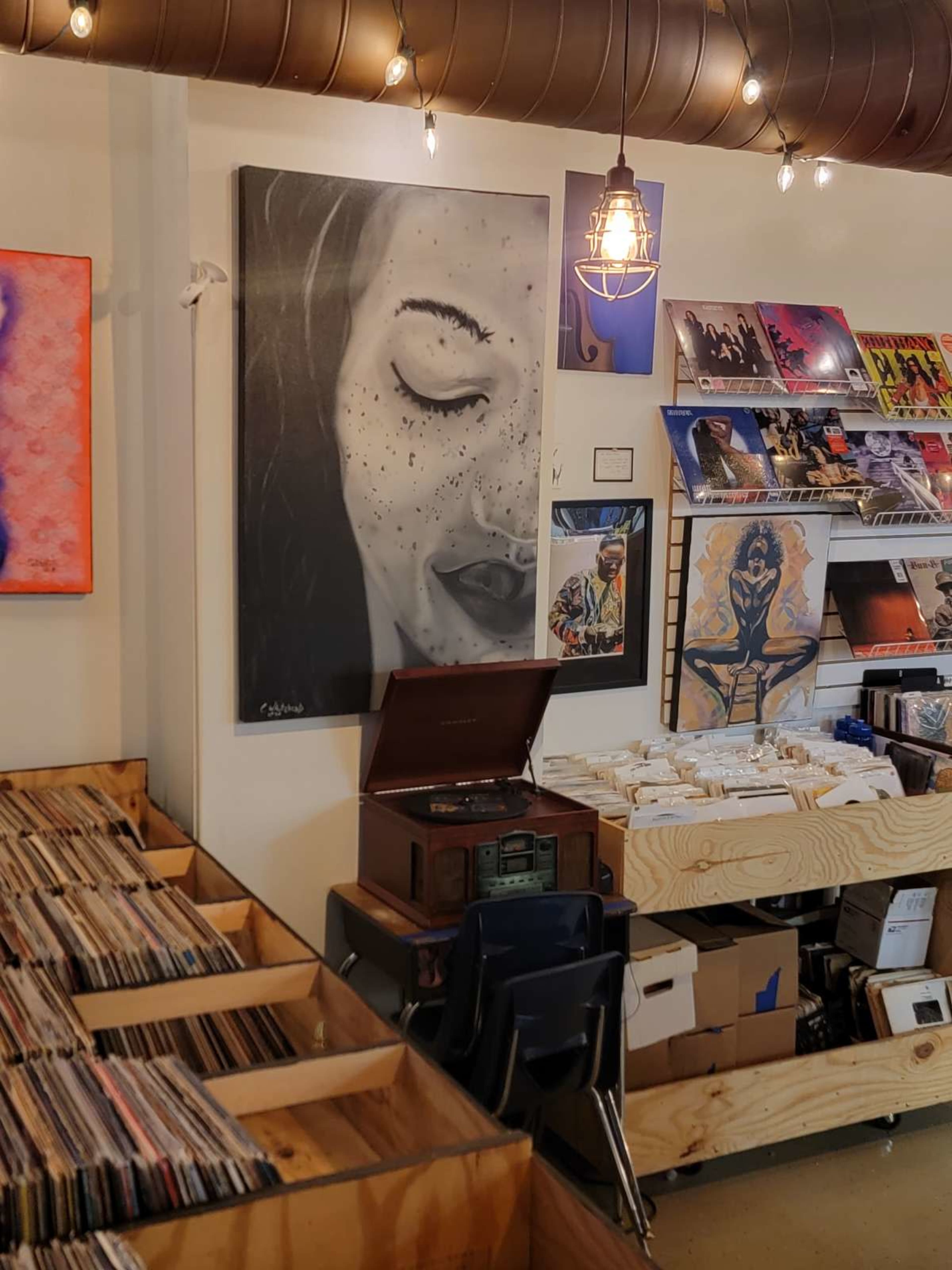 The image shows a record store with wooden shelves filled with vinyl records, a vintage turntable on display, and artwork of a woman's face hanging on the wall.