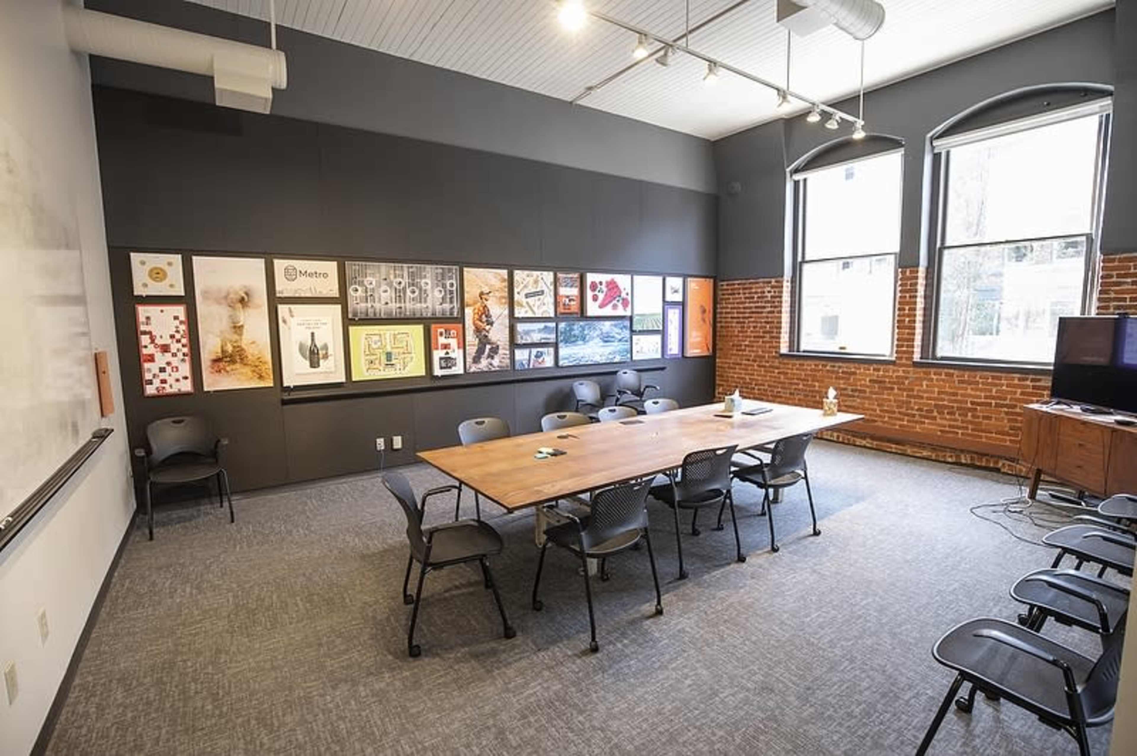 A conference room features a long wooden table surrounded by chairs, with artwork displayed on the walls and large windows allowing natural light.