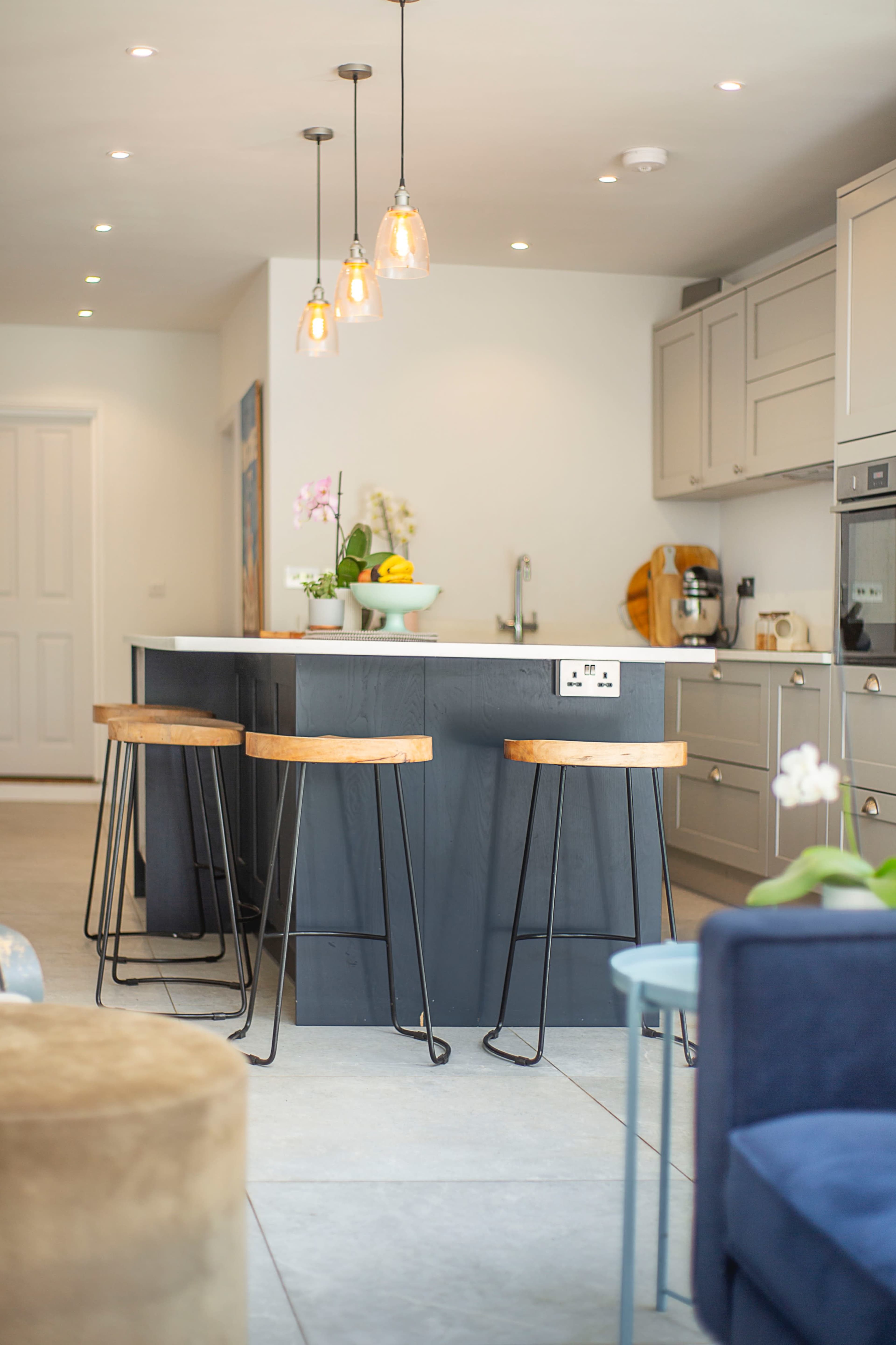 The image shows a modern kitchen with a central island, three wooden bar stools, and pendant lights hanging above.