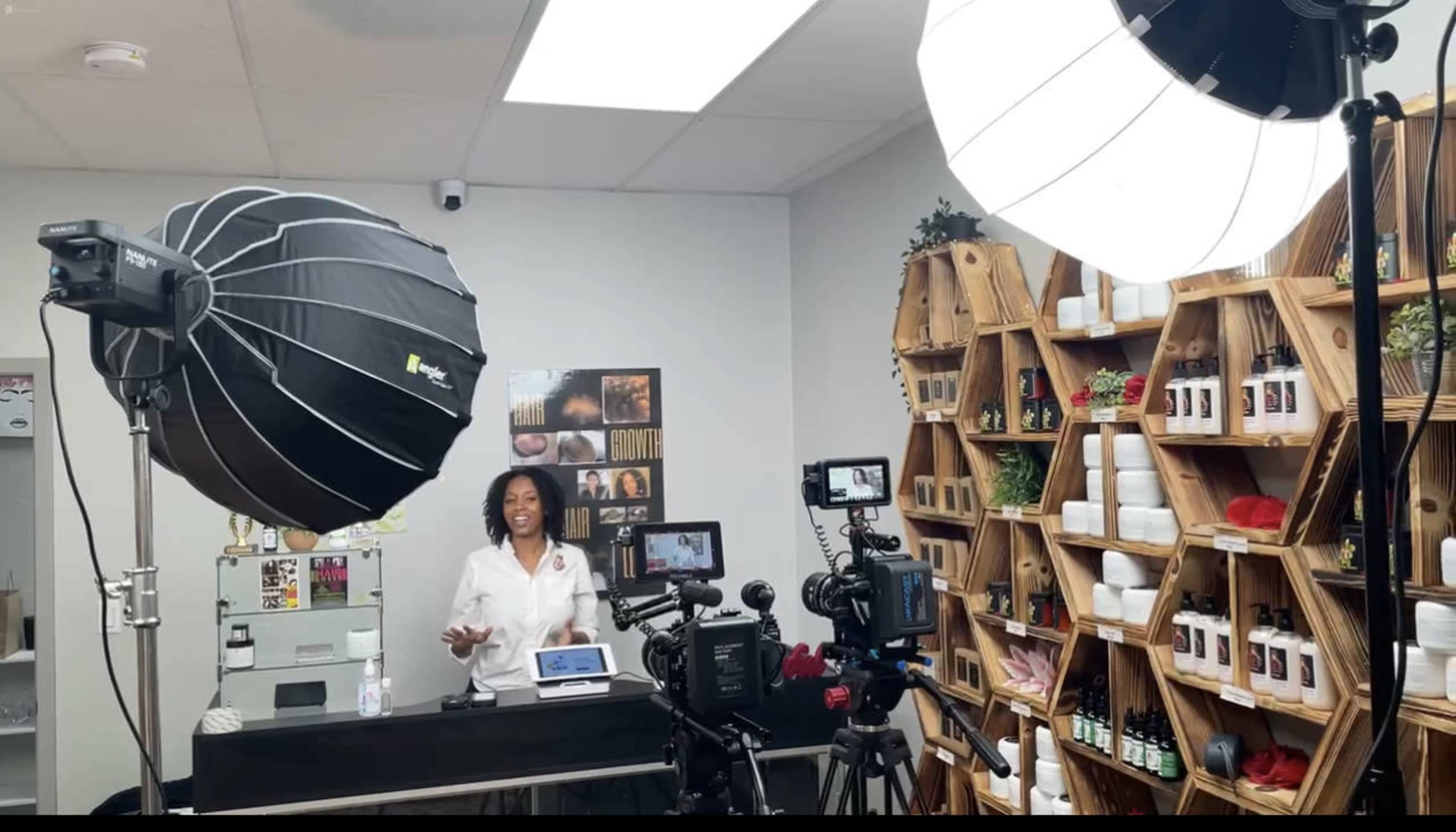 A woman stands behind a counter in a well-arranged studio filled with beauty products, flanked by professional cameras and lighting equipment.