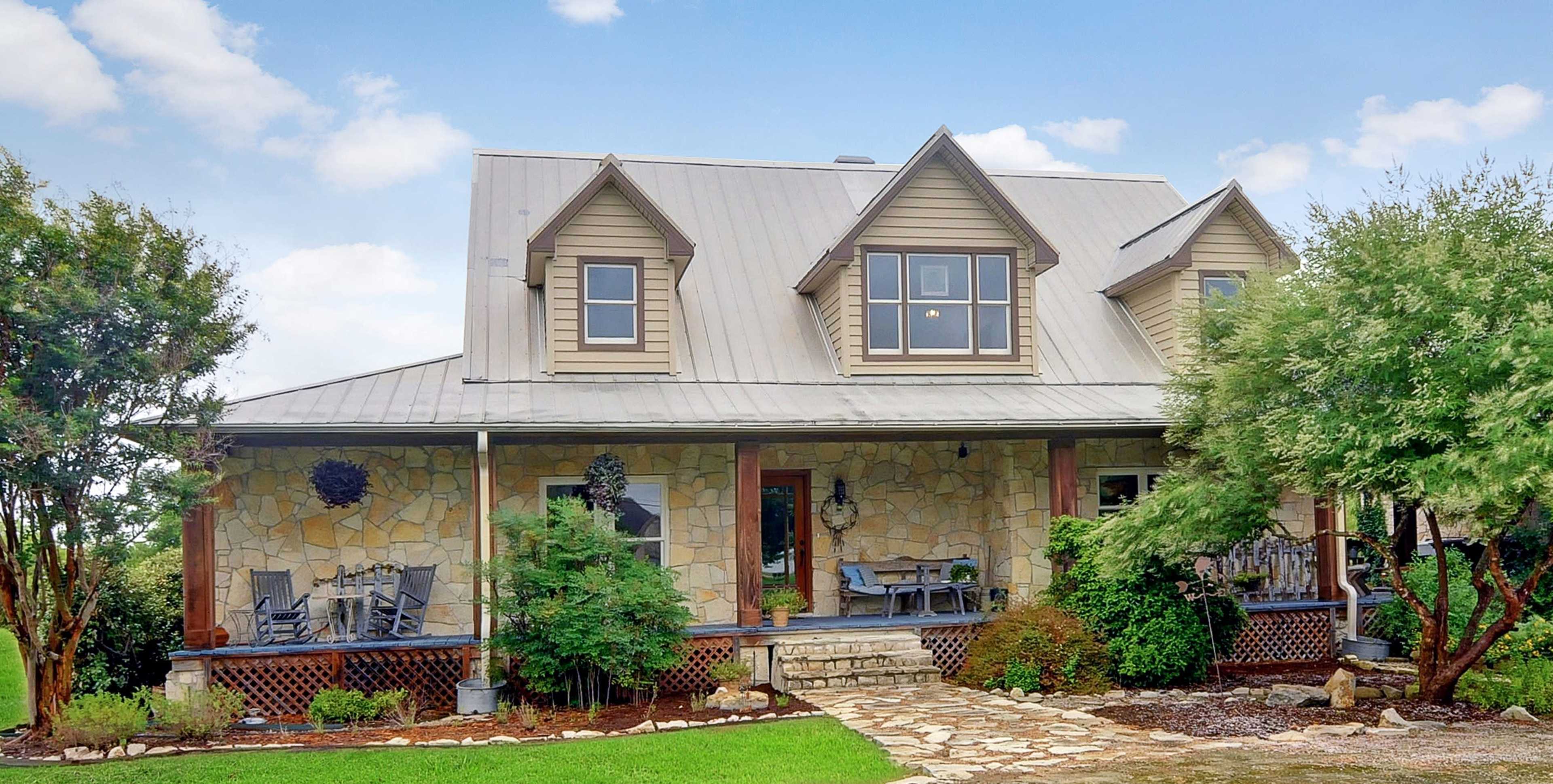 A two-story house with a stone facade, a metal roof, and a welcoming porch surrounded by greenery and landscaped grounds.