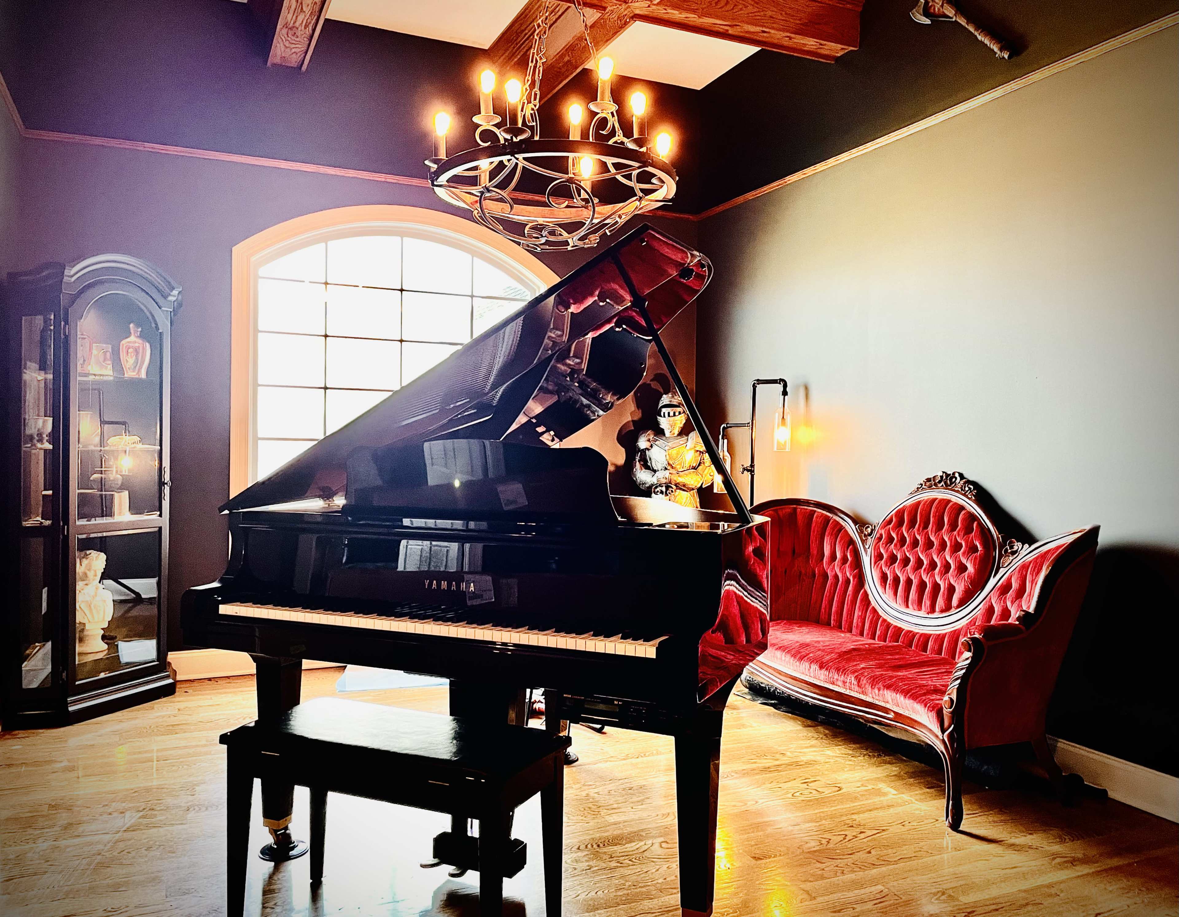 The scene features a grand piano beside a vintage red velvet couch in a dimly lit room with dark walls and a chandelier overhead.