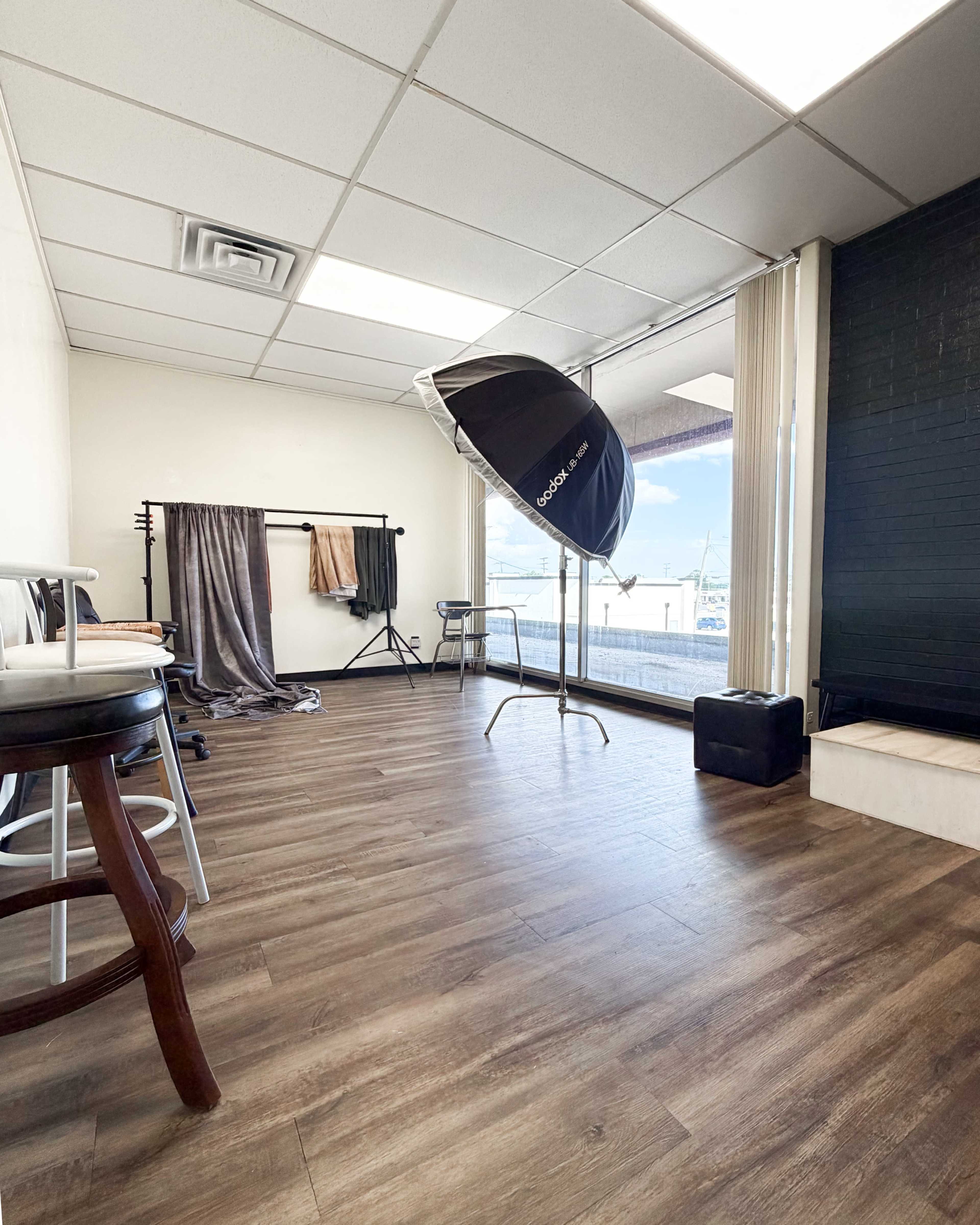 The image shows a minimalist studio space with a large window, a backdrop stand draped with fabric, a light reflector, and a few pieces of furniture.