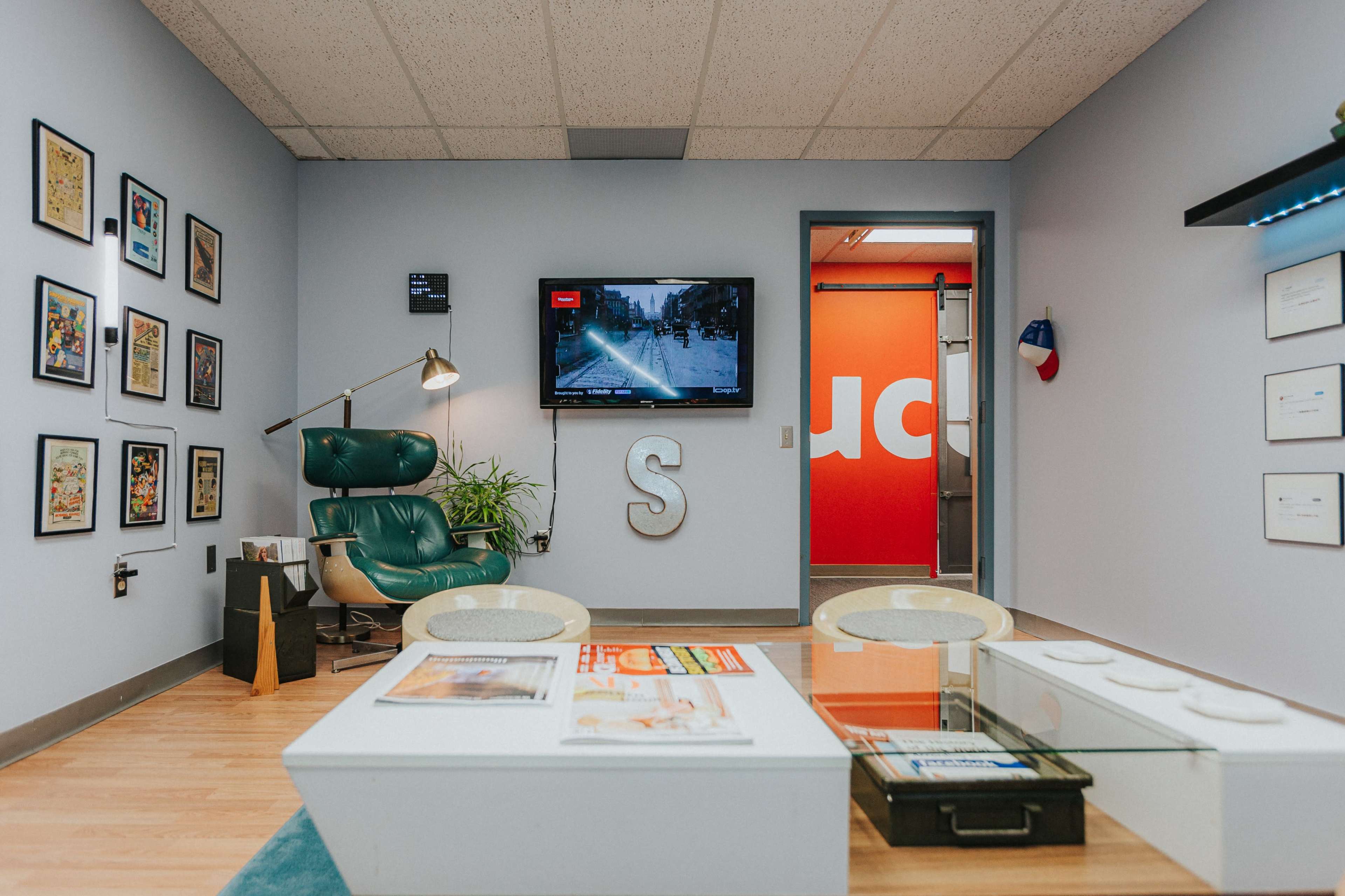A modern office space featuring a green lounge chair, a large TV on the wall displaying a street view, and decorative framed artworks on the walls.