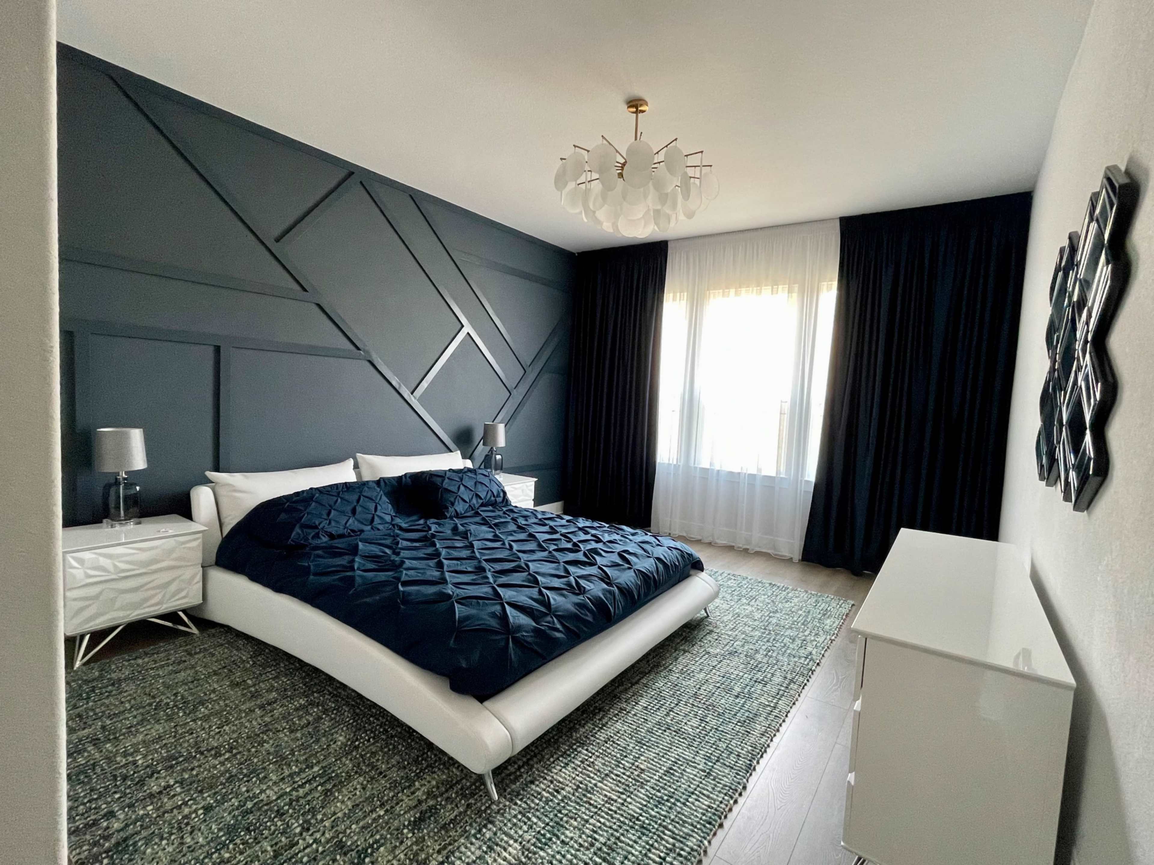 The image shows a modern bedroom with a large bed, two bedside tables with lamps, a white dresser, and a window with sheer curtains.