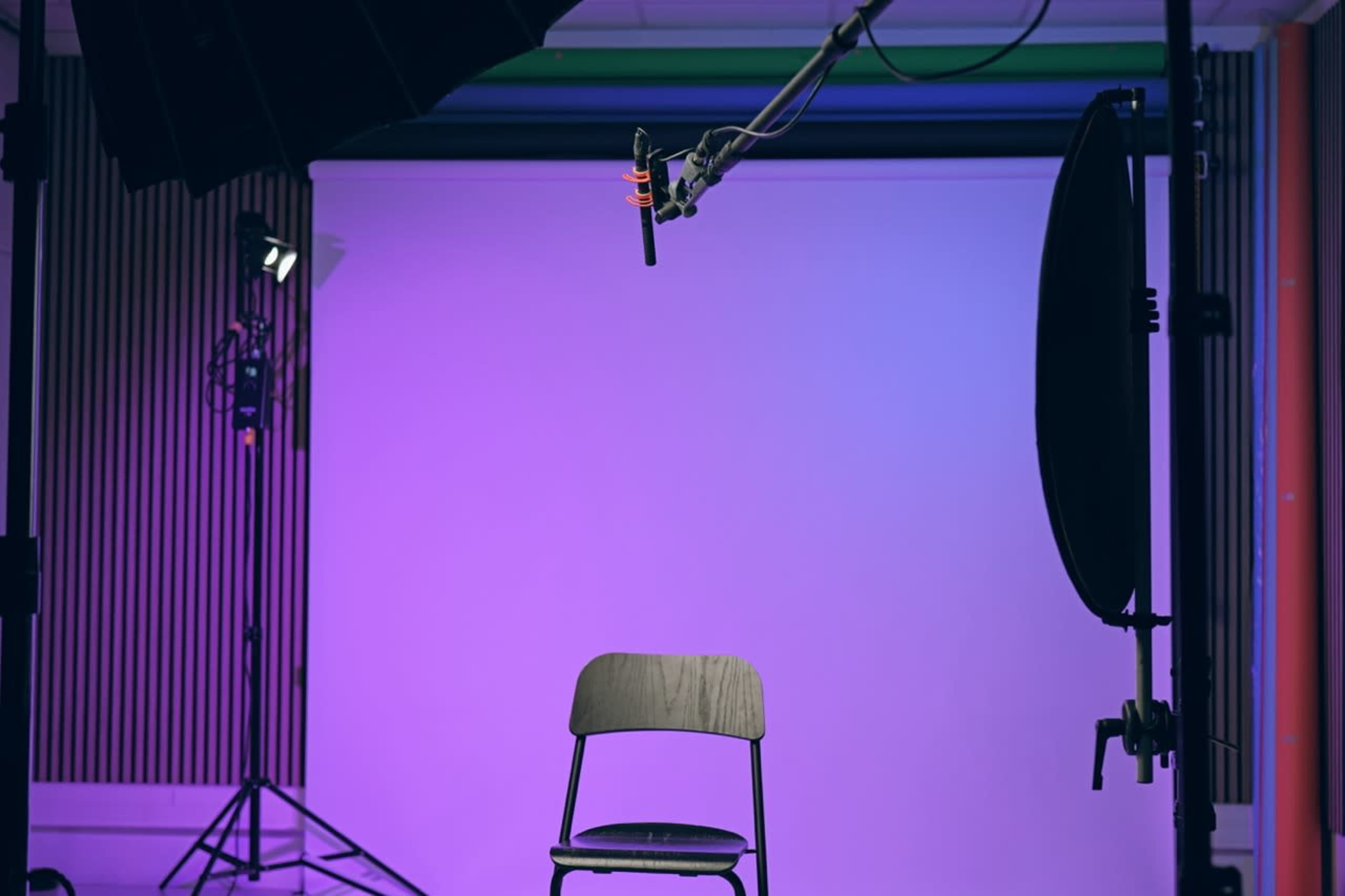 A wooden chair is positioned in front of a soft-lit backdrop with purple hues, surrounded by studio lights and a microphone on an adjustable arm.