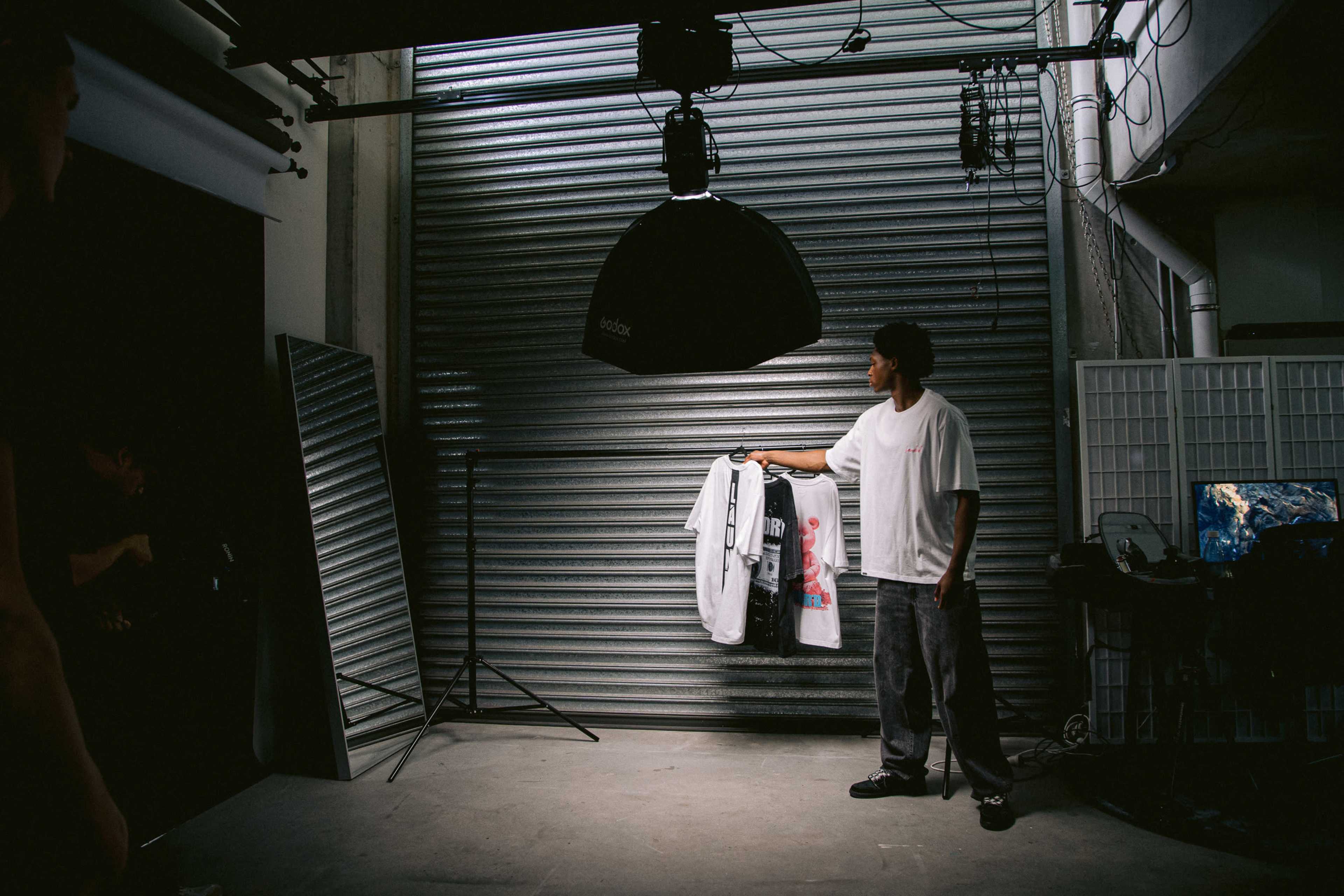 A person in a white shirt holds up a t-shirt in front of a backdrop with a metal shutter in a photography studio.