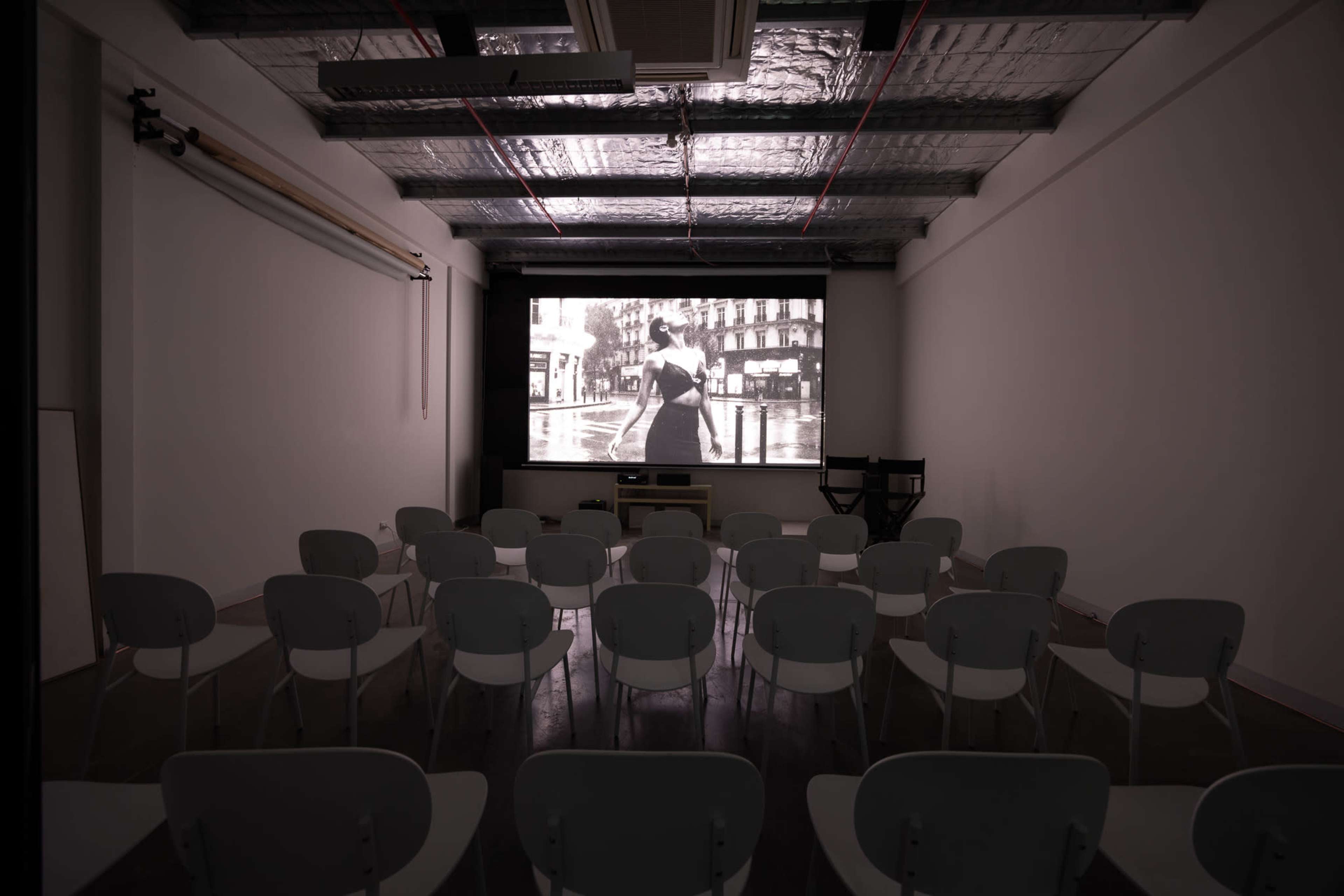 The image shows a sparsely furnished room with rows of white chairs facing a large projection screen displaying a black-and-white film.