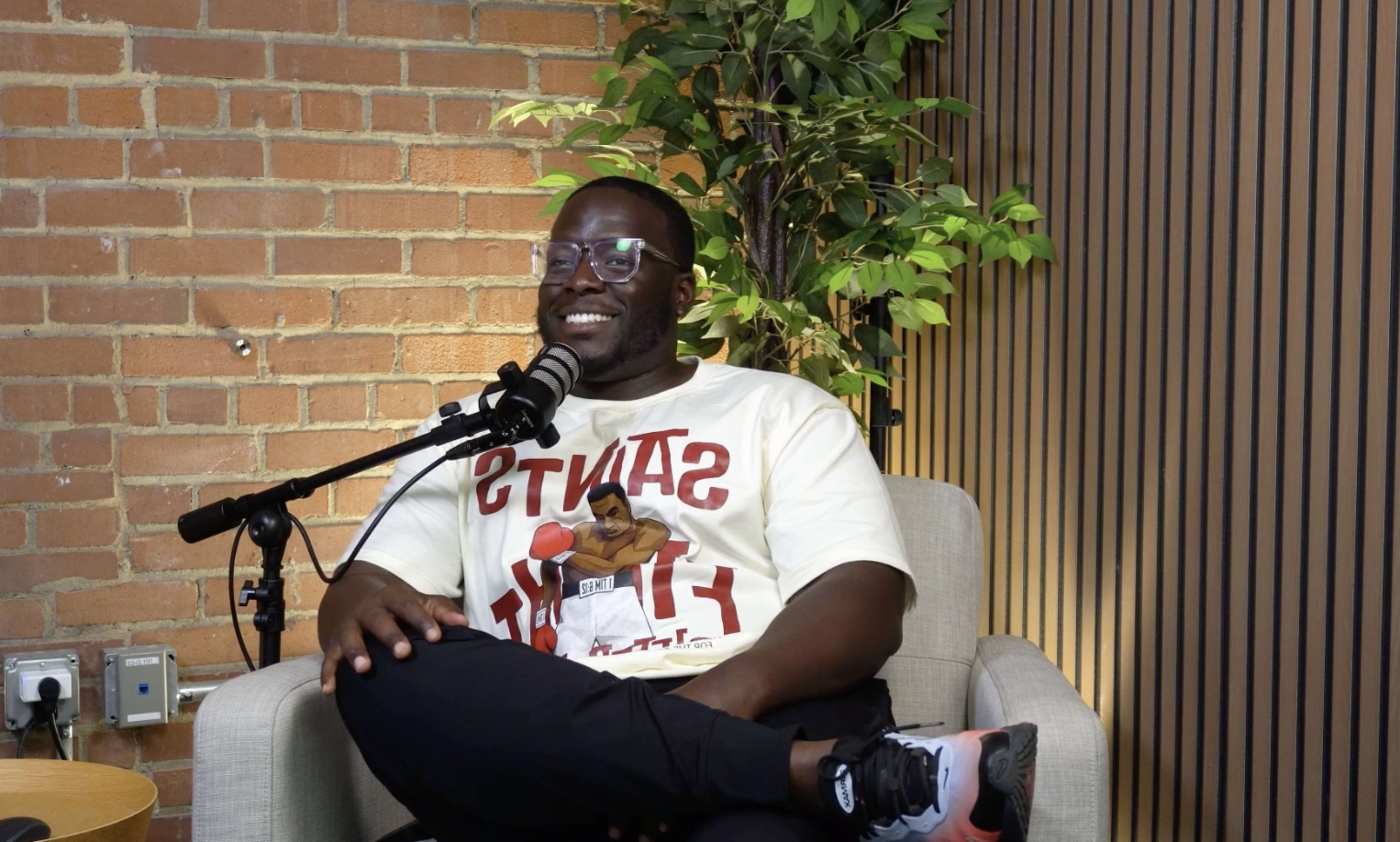 A man sits in a chair with a microphone in front of him, wearing a t-shirt with the words "SAINTS" and "HIT," while smiling and looking at the camera.