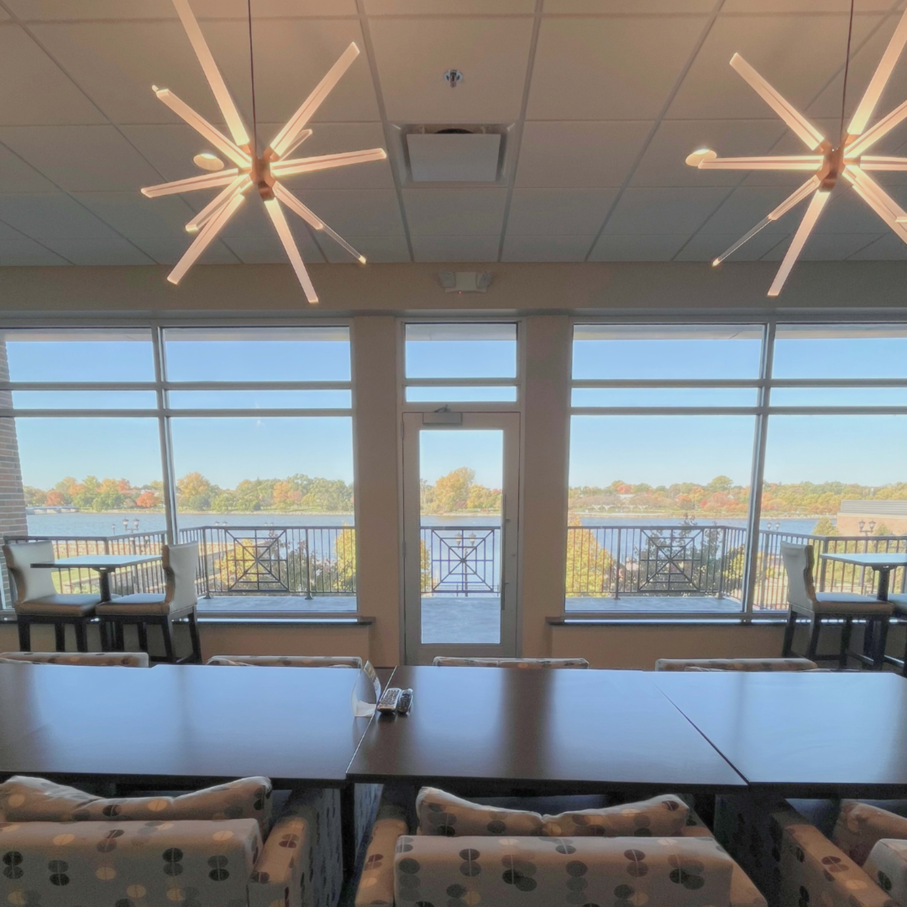 The image shows a spacious room with modern lighting fixtures and large windows overlooking a body of water and trees in the background.