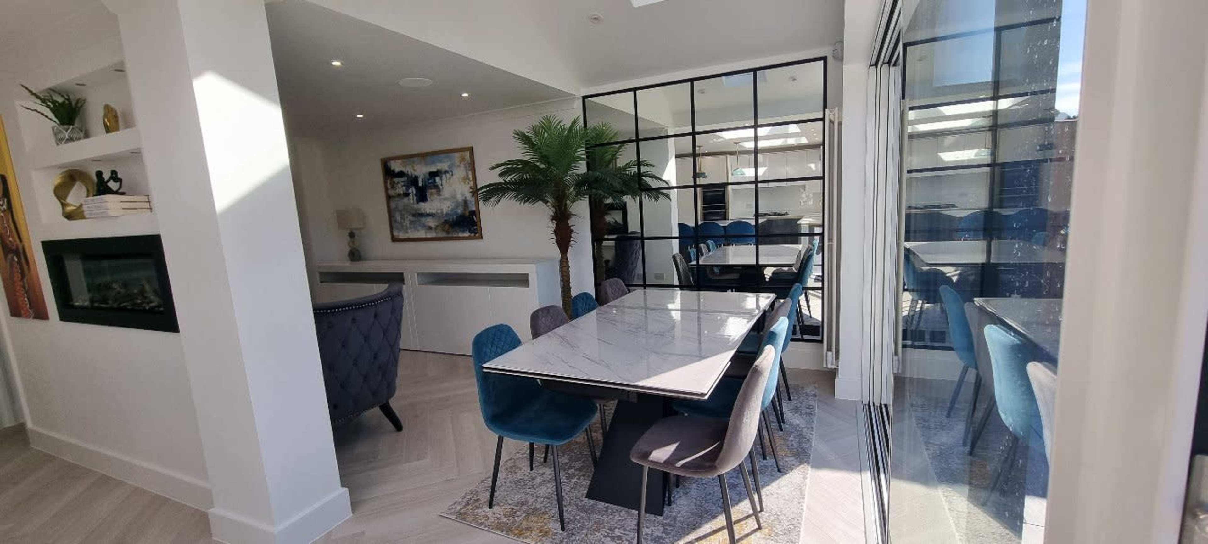 A modern dining area features a long table surrounded by various upholstered chairs, with large glass doors leading to an outdoor space and a decorative palm in the corner.
