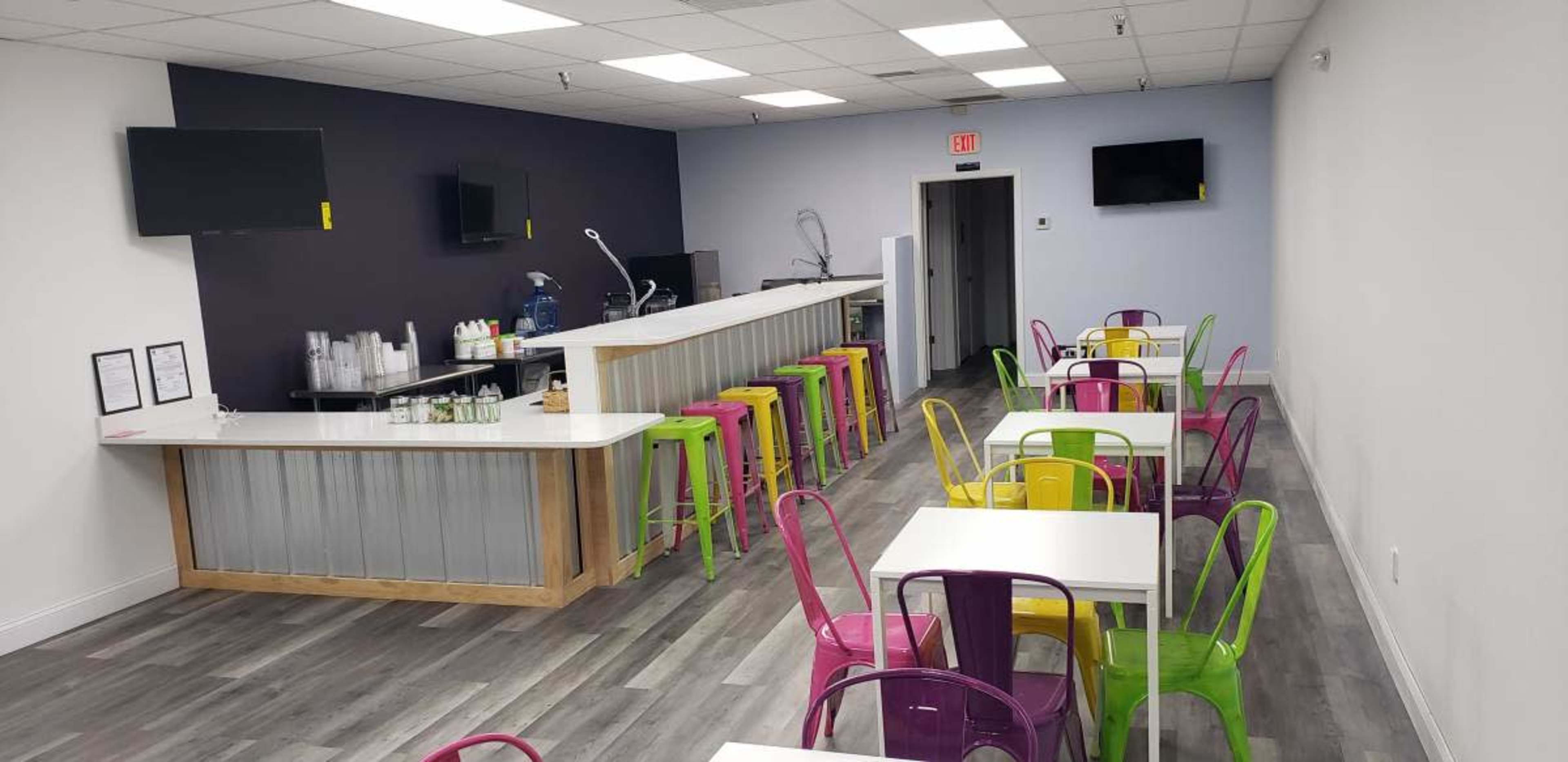 The image shows a modern, brightly colored café with a long bar area and multiple tables and chairs arranged throughout the space.