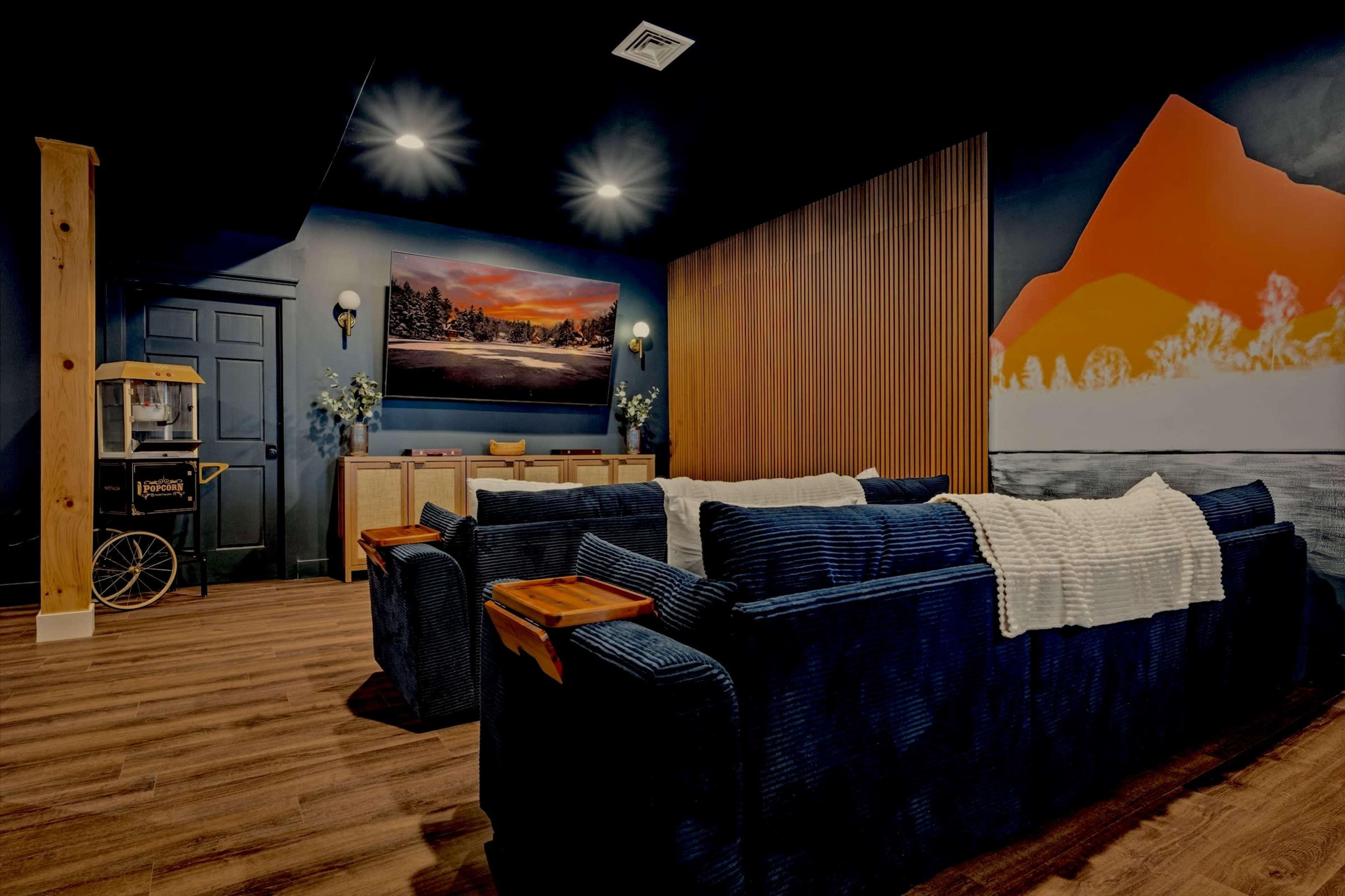 The image shows a cozy indoor lounge area featuring navy blue couches, a wooden accent wall, and a large mural depicting a mountain landscape.