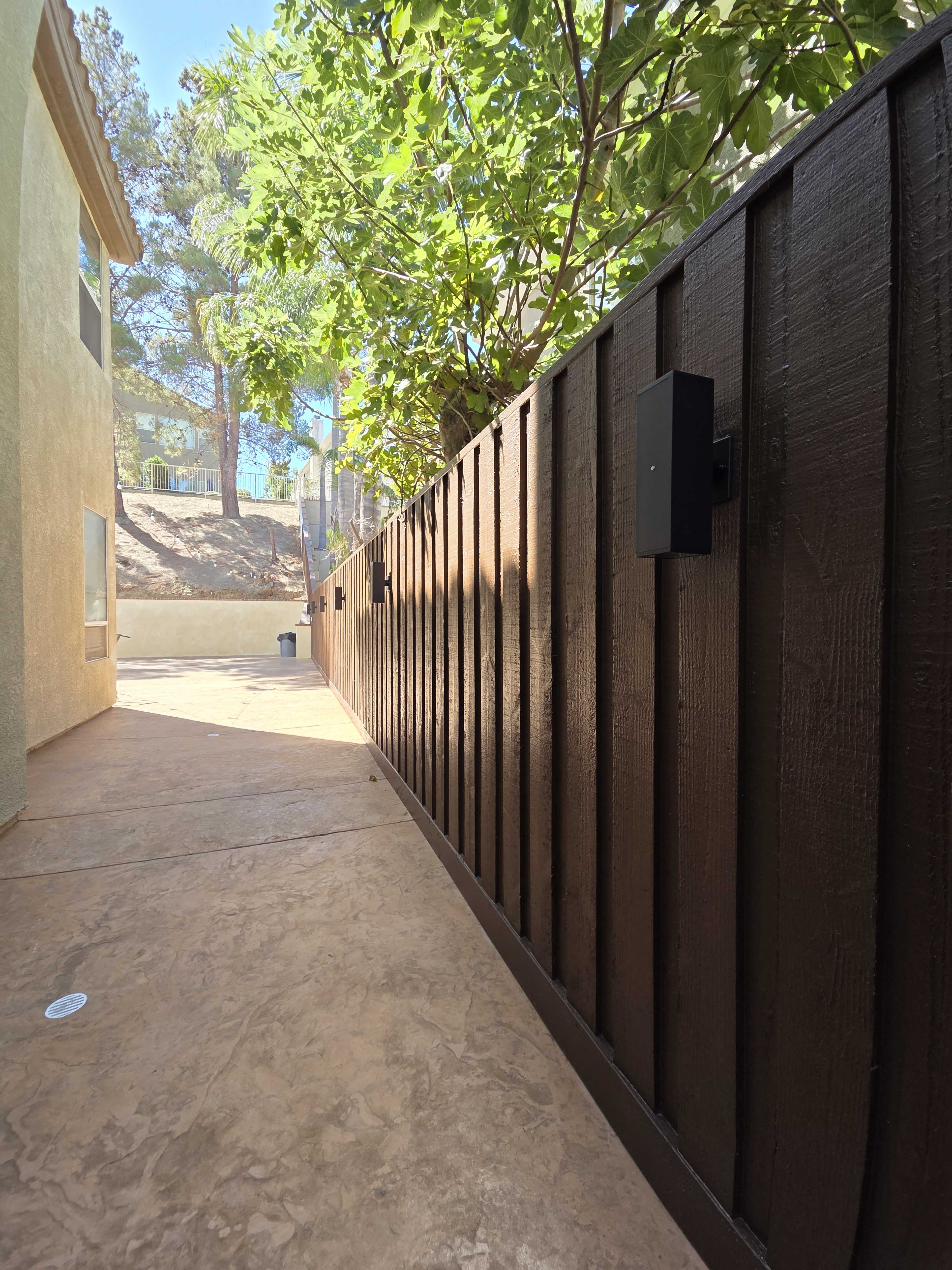 The image shows a narrow walkway beside a wooden fence with black light fixtures, surrounded by trees and a building on one side.