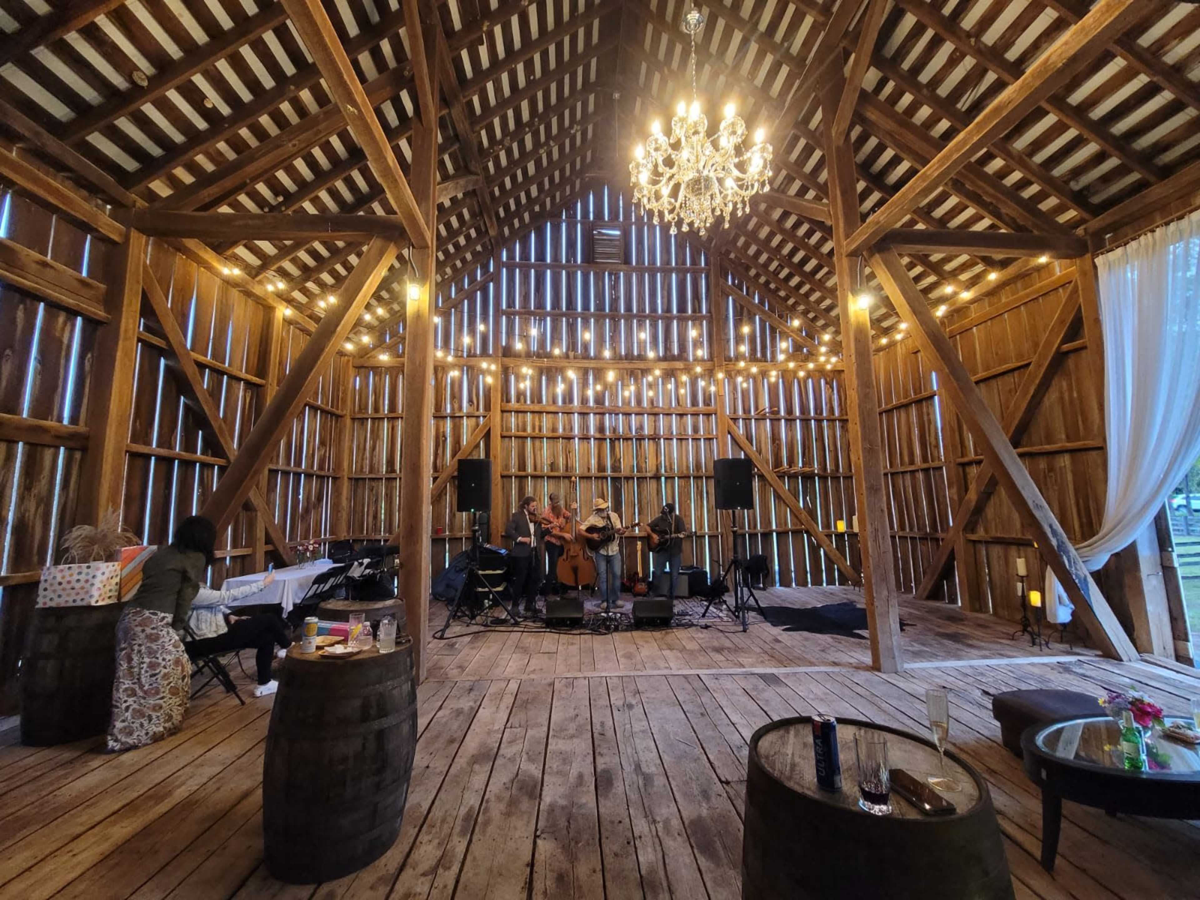A wooden barn interior features a live band performing under a chandelier, with string lights illuminating the space.