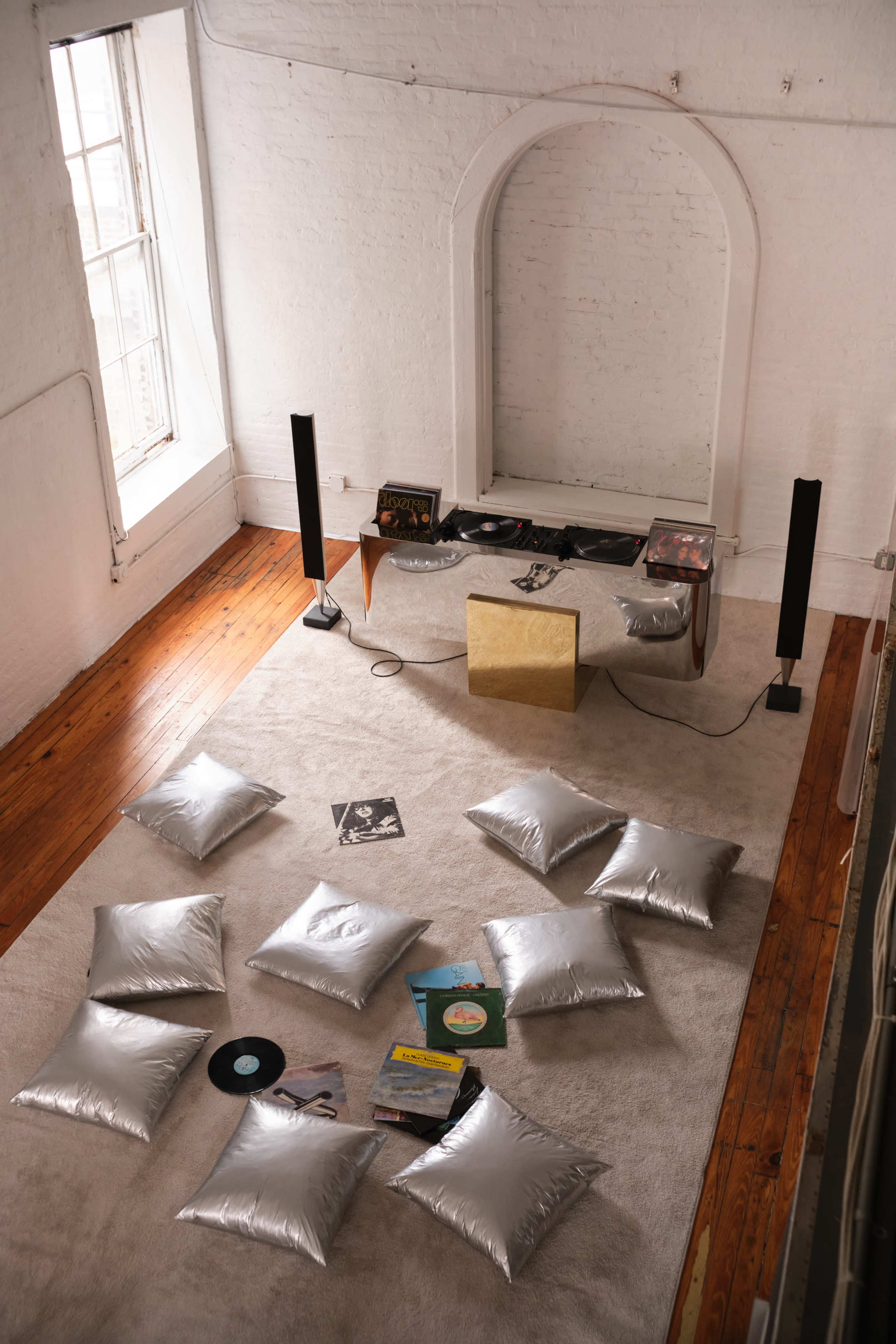 The image shows a spacious room with a light-colored carpet, scattered silver pillows, a small table with a record player, and several vinyl records on the floor.