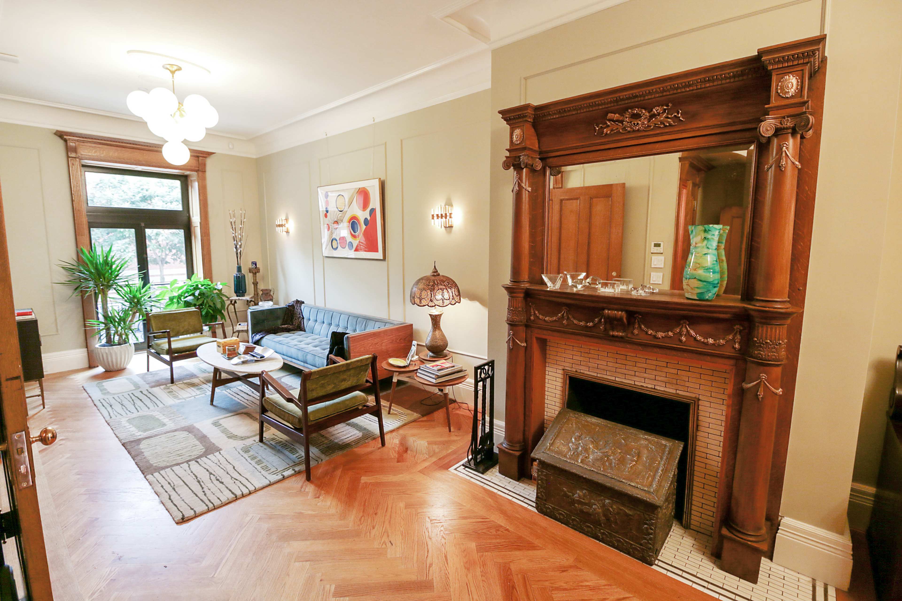 A living room with a vintage wooden fireplace, a blue sofa, and a round coffee table surrounded by chairs, accented by plants and art on the walls.