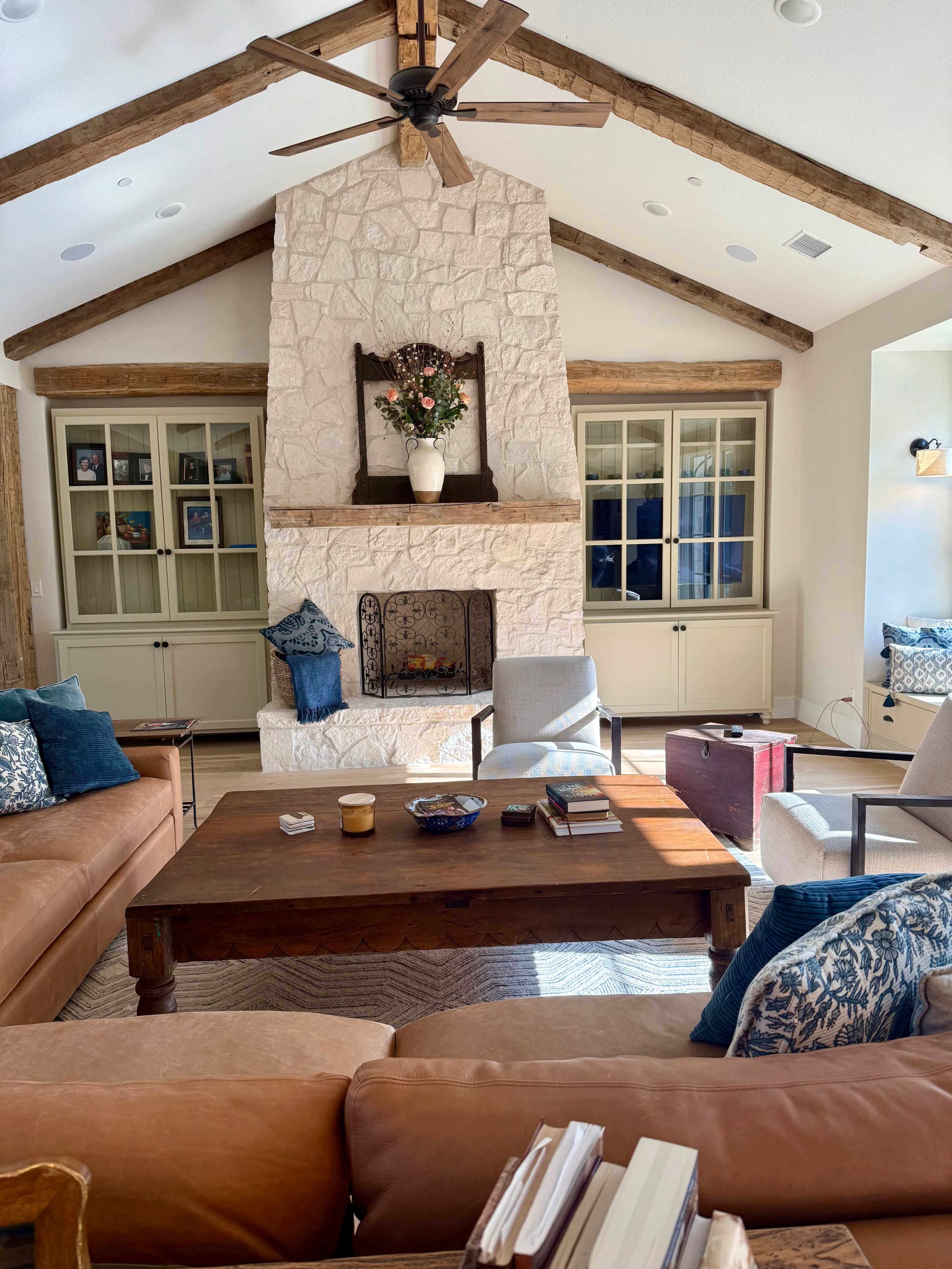 The living room features a stone fireplace with a wooden mantle, surrounded by two brown sofas, a wooden coffee table, and a ceiling fan above.