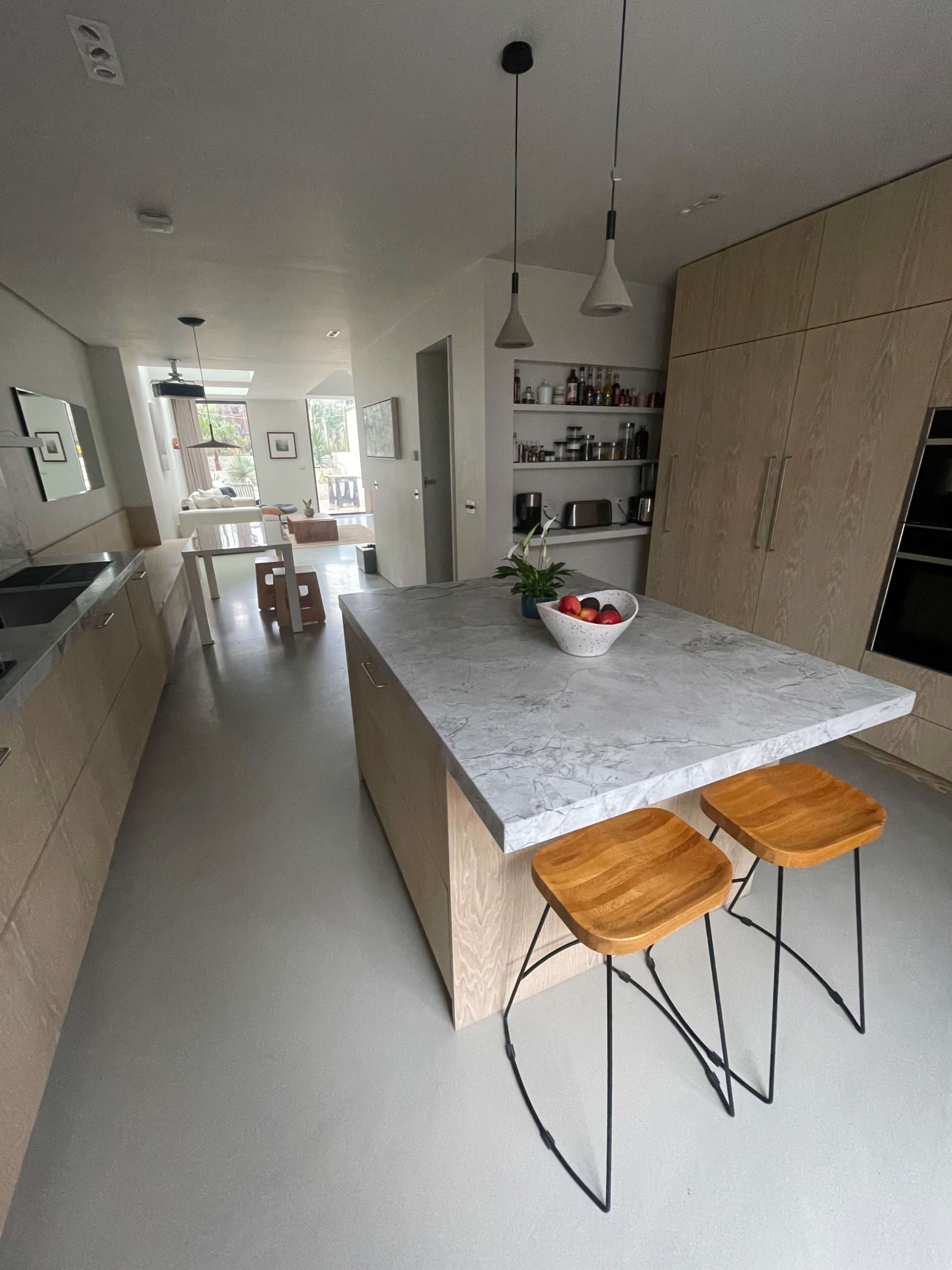 The image shows a modern kitchen with a central marble-topped island, two wooden bar stools, and shelves displaying kitchen items.