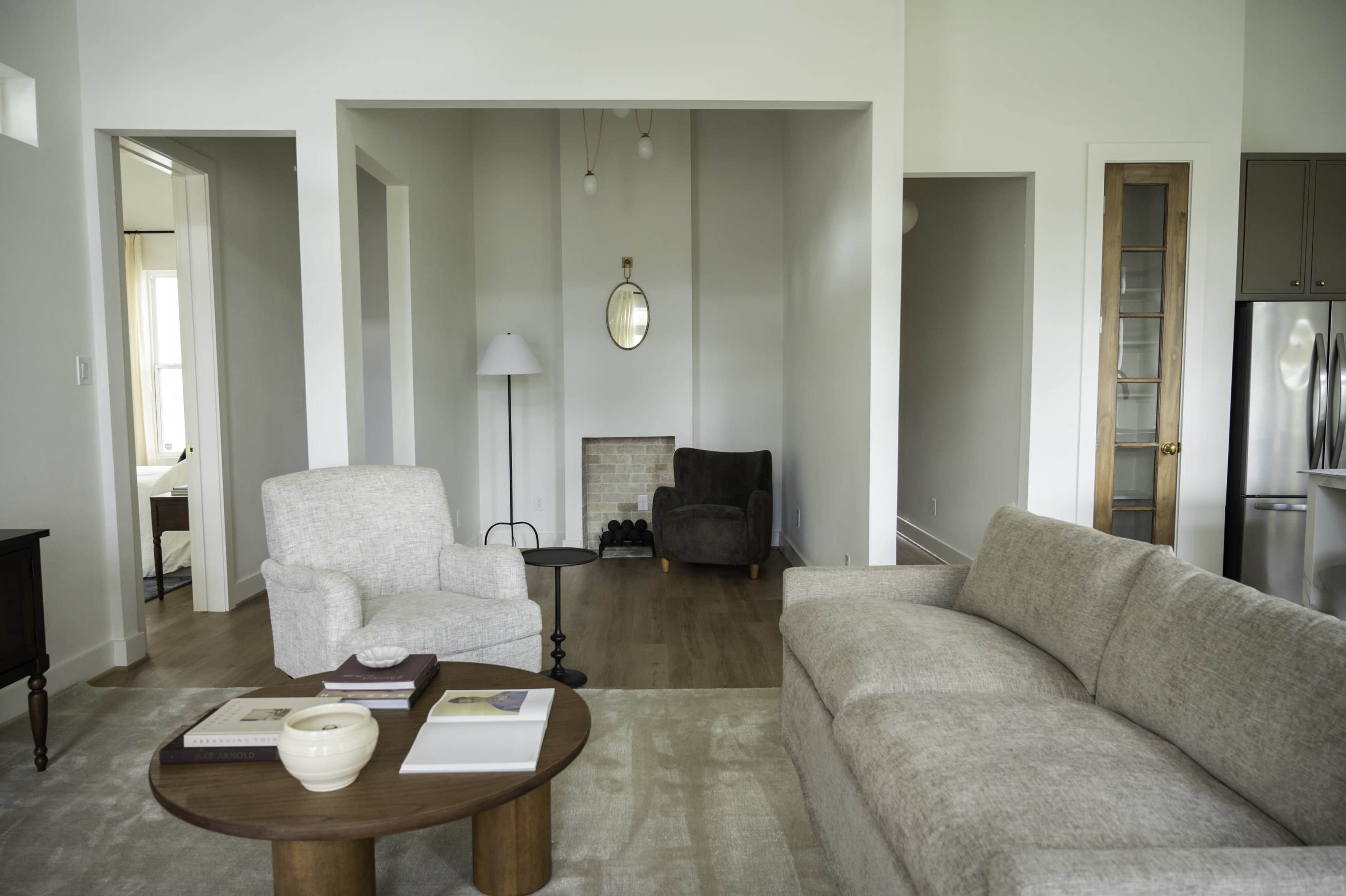 The image shows a modern living room with beige furniture, a round coffee table, and an open layout connecting different areas of the space.
