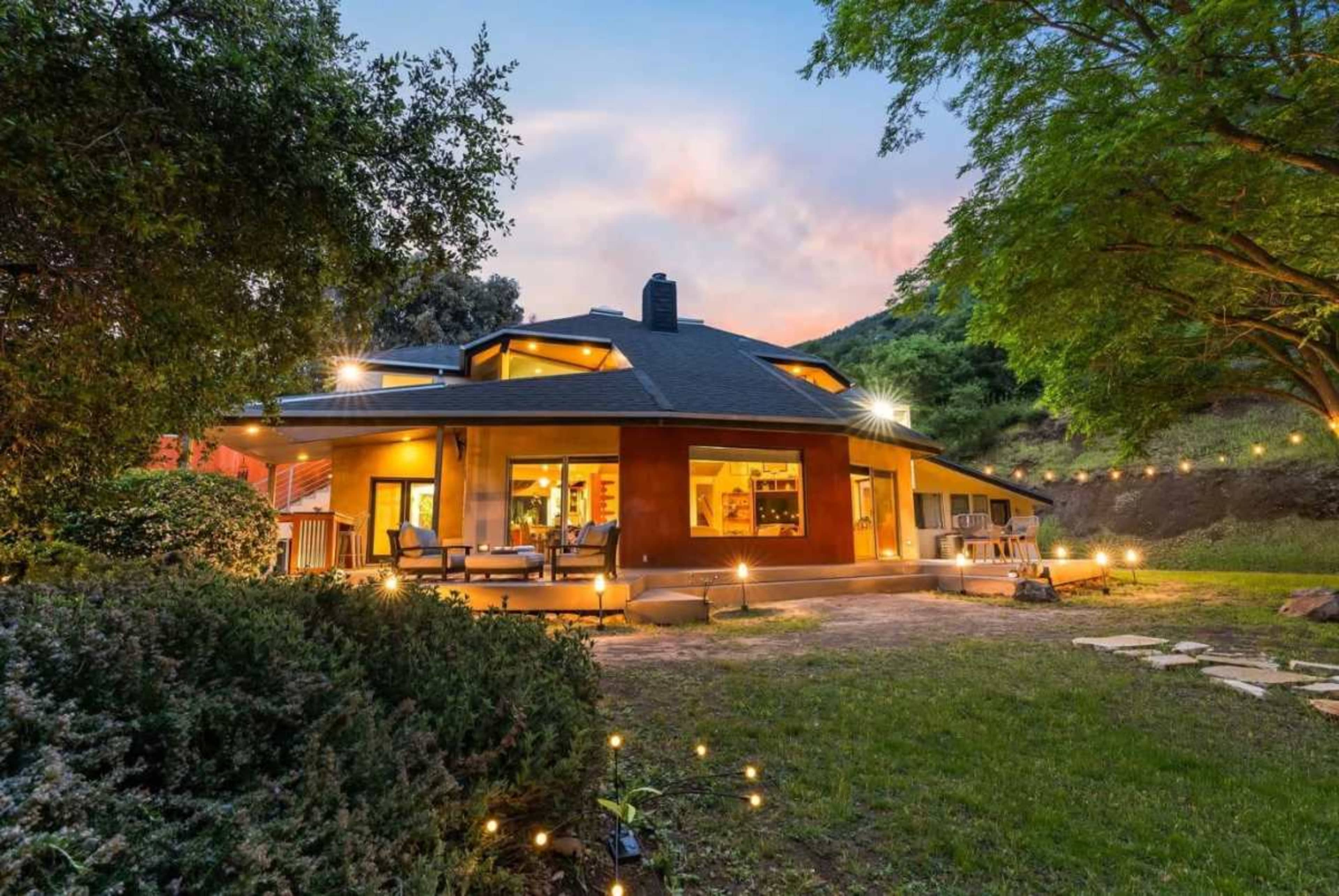 A modern house with a rounded design is situated on a grassy area, surrounded by trees and outdoor lighting.