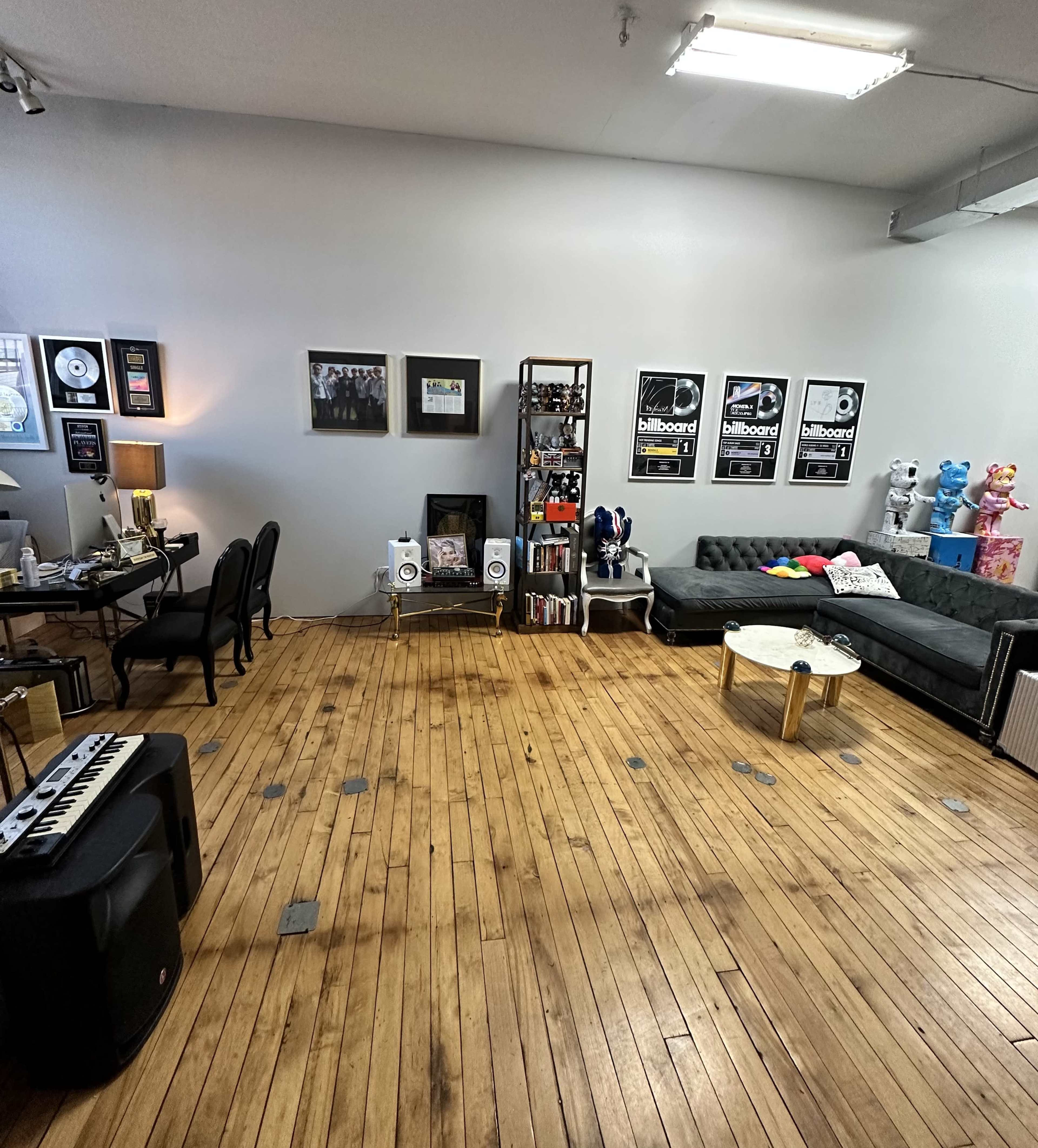 The image shows a spacious room with wooden floors, featuring a seating area with a couch and colorful decorations, along with a workspace that includes a desk and musical equipment.