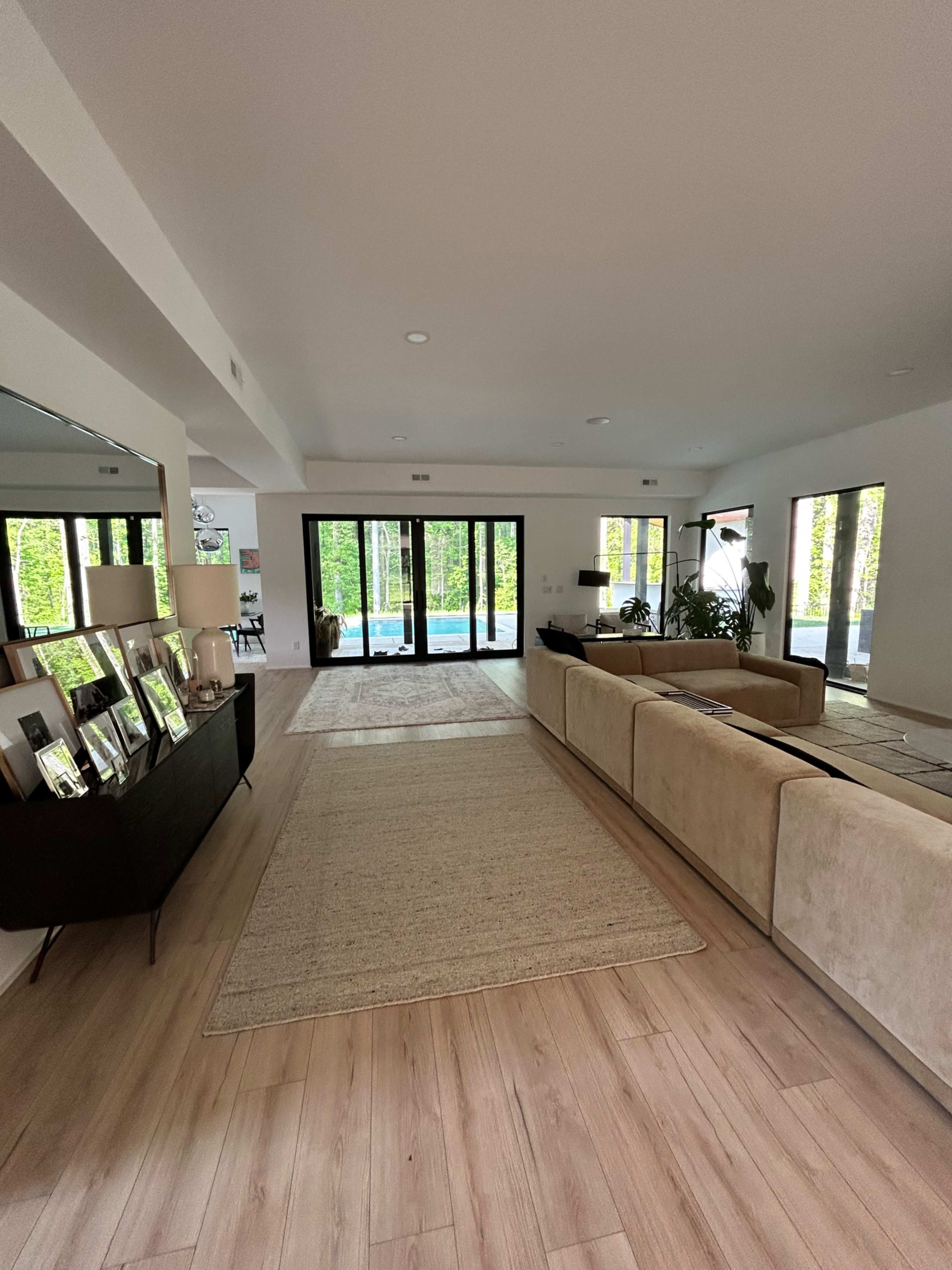 The image shows a modern, open-concept living space with a beige sectional sofa, a large mirror, and sliding glass doors leading to a pool area surrounded by greenery.